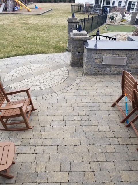 Stone patio with a circular design, outdoor kitchen, lawn, and chairs.