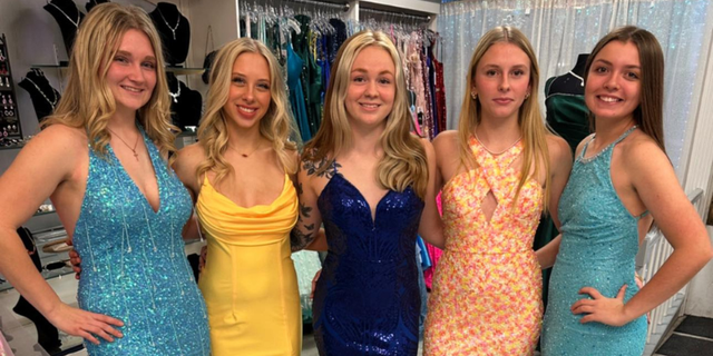 Five young women in sparkly formal dresses posing together indoors, smiling at the camera.
