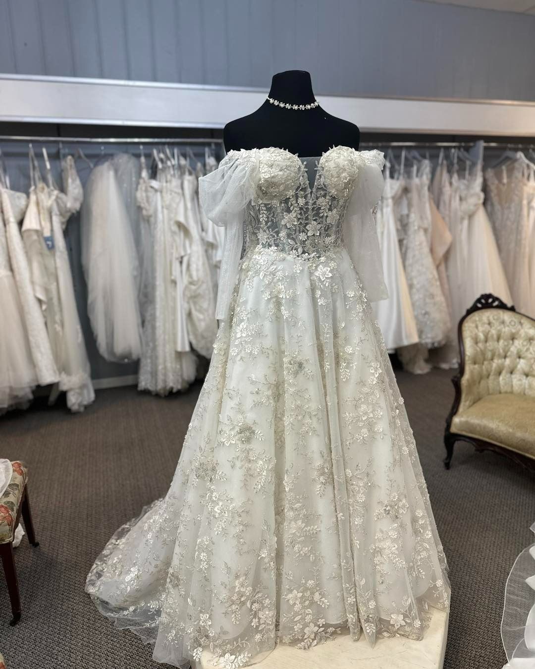 White beaded wedding gown on mannequin in bridal shop, with dresses hanging in the background