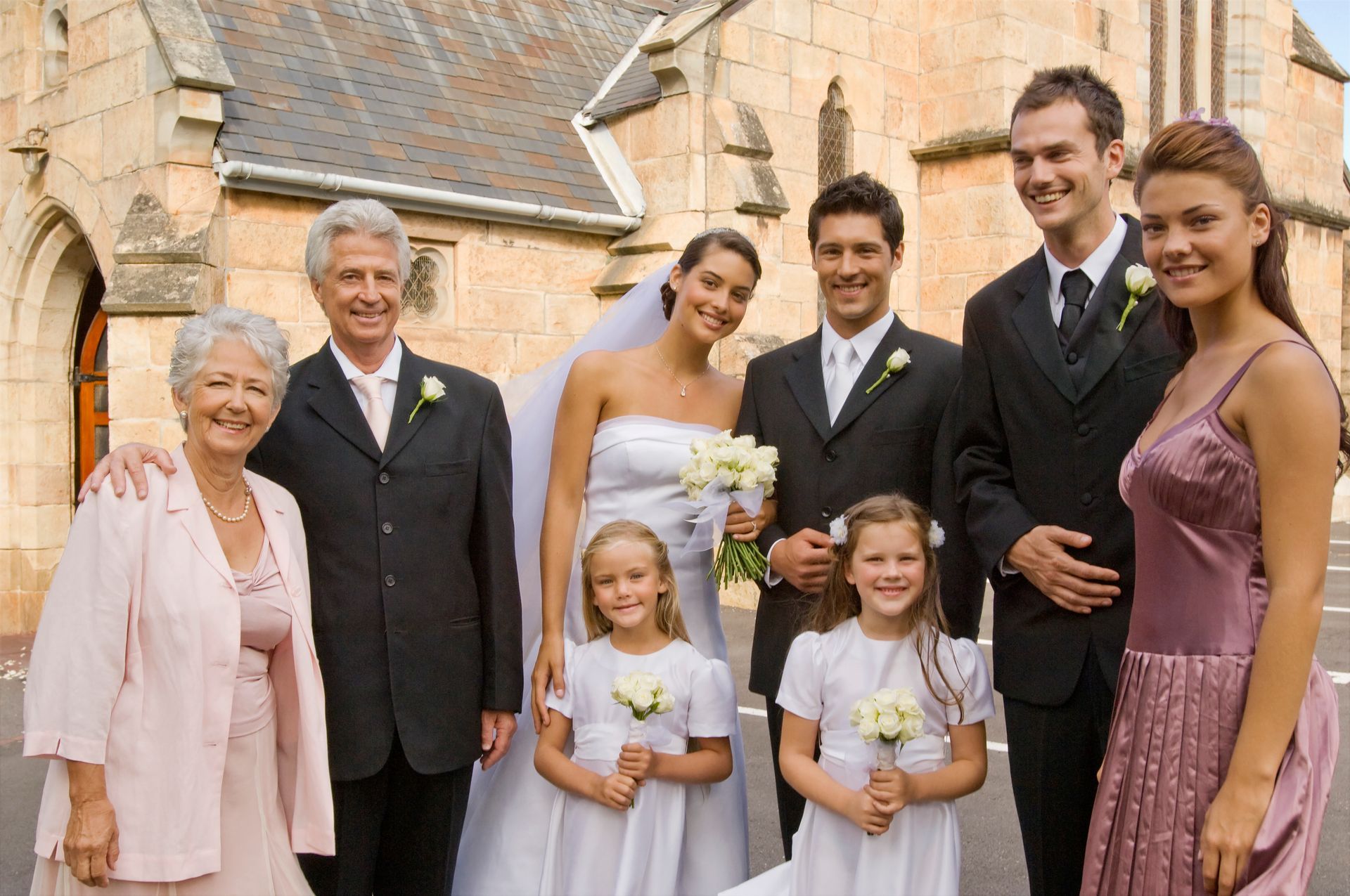 Wedding party posing outside a stone building, with bride, groom, children, and family members smiling.