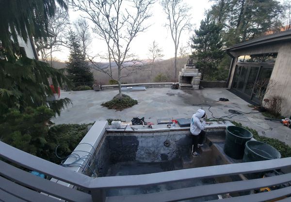 Person cleaning an outdoor hot tub on a patio with a backyard and trees beyond