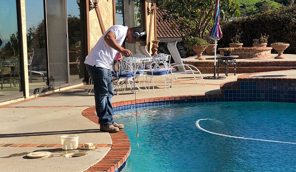 Person rinsing off by a swimming pool with a garden hose on a sunny patio.