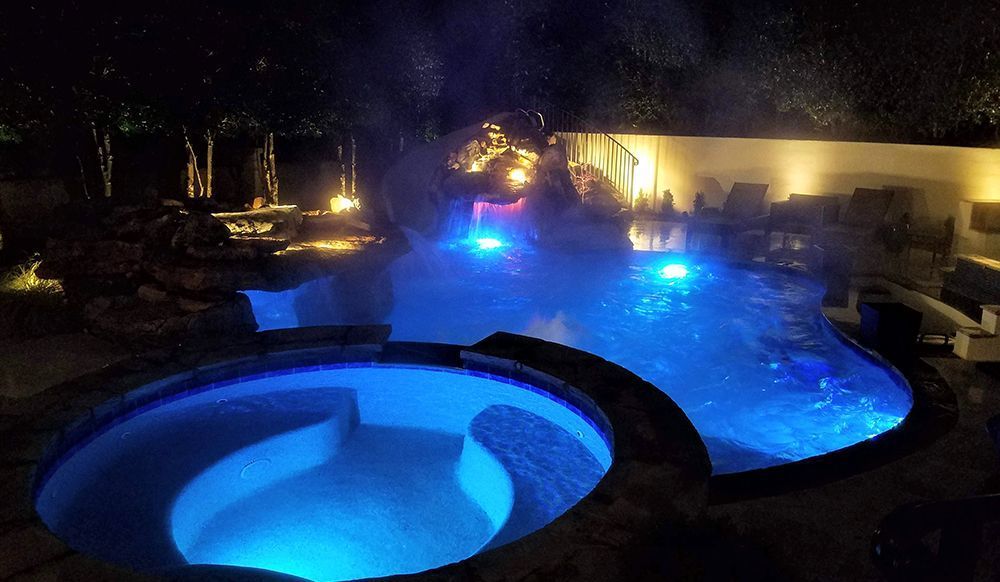Nighttime backyard pool and hot tub glowing blue with warm garden lights and mist