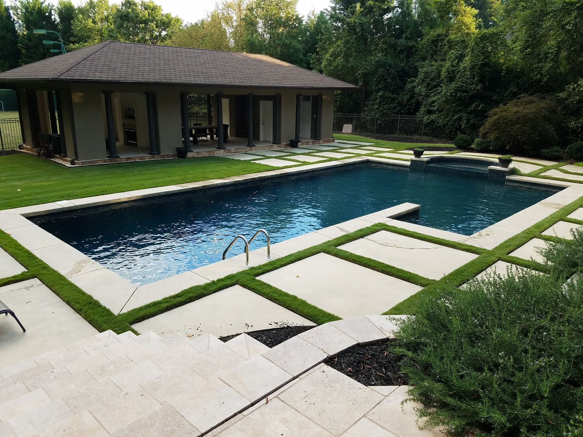 Modern backyard with a rectangular pool, stone patio, and a house surrounded by trees