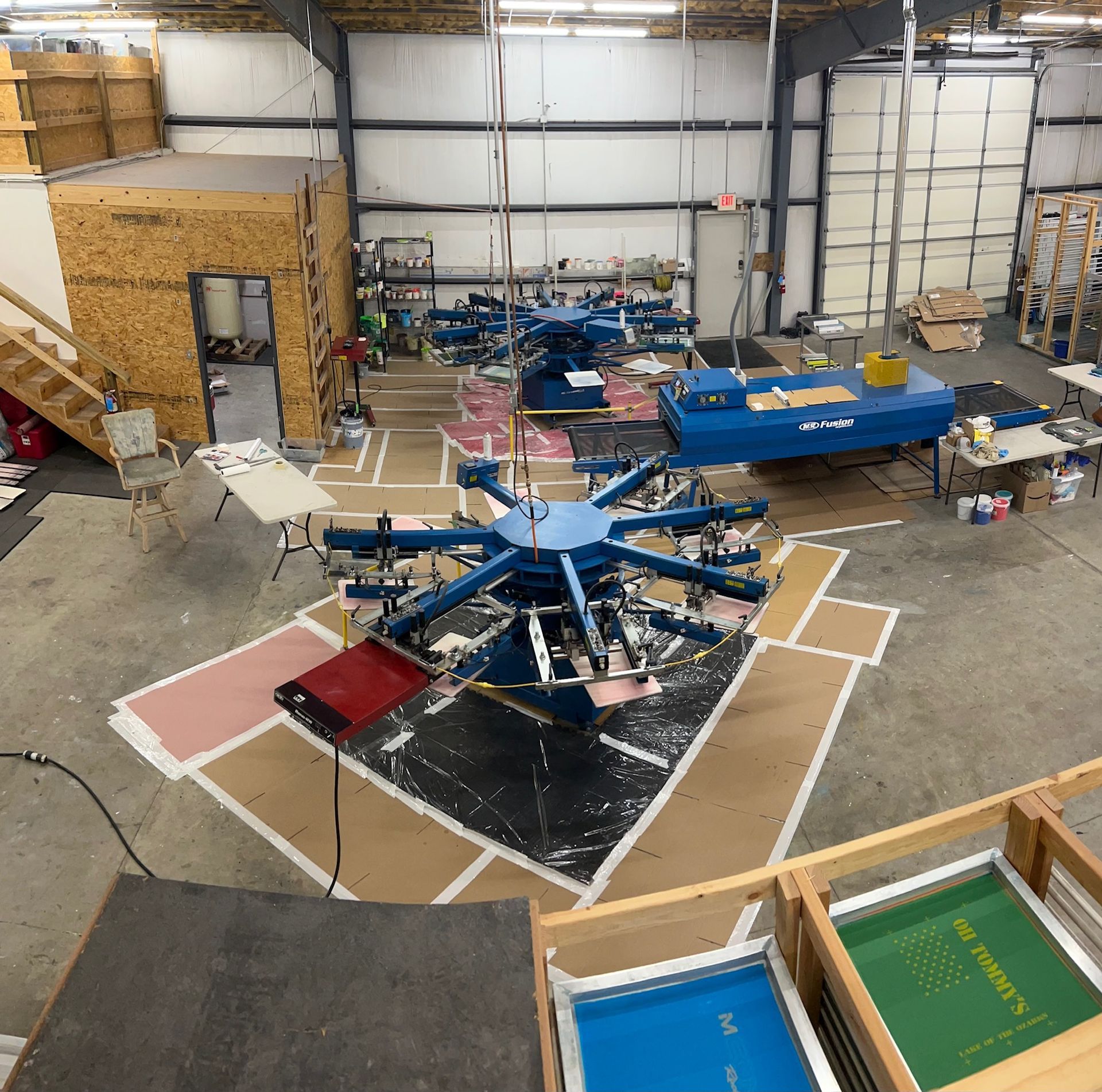 Screen printing shop with blue printing machines, wooden screens, and supplies in a large warehouse.