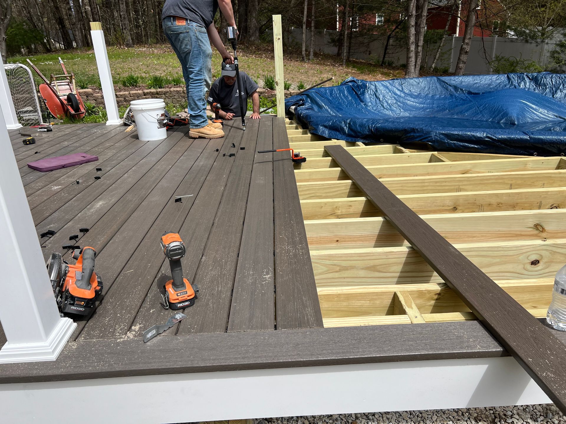 Men building a deck with gray composite boards, drilling, with wood framing visible.