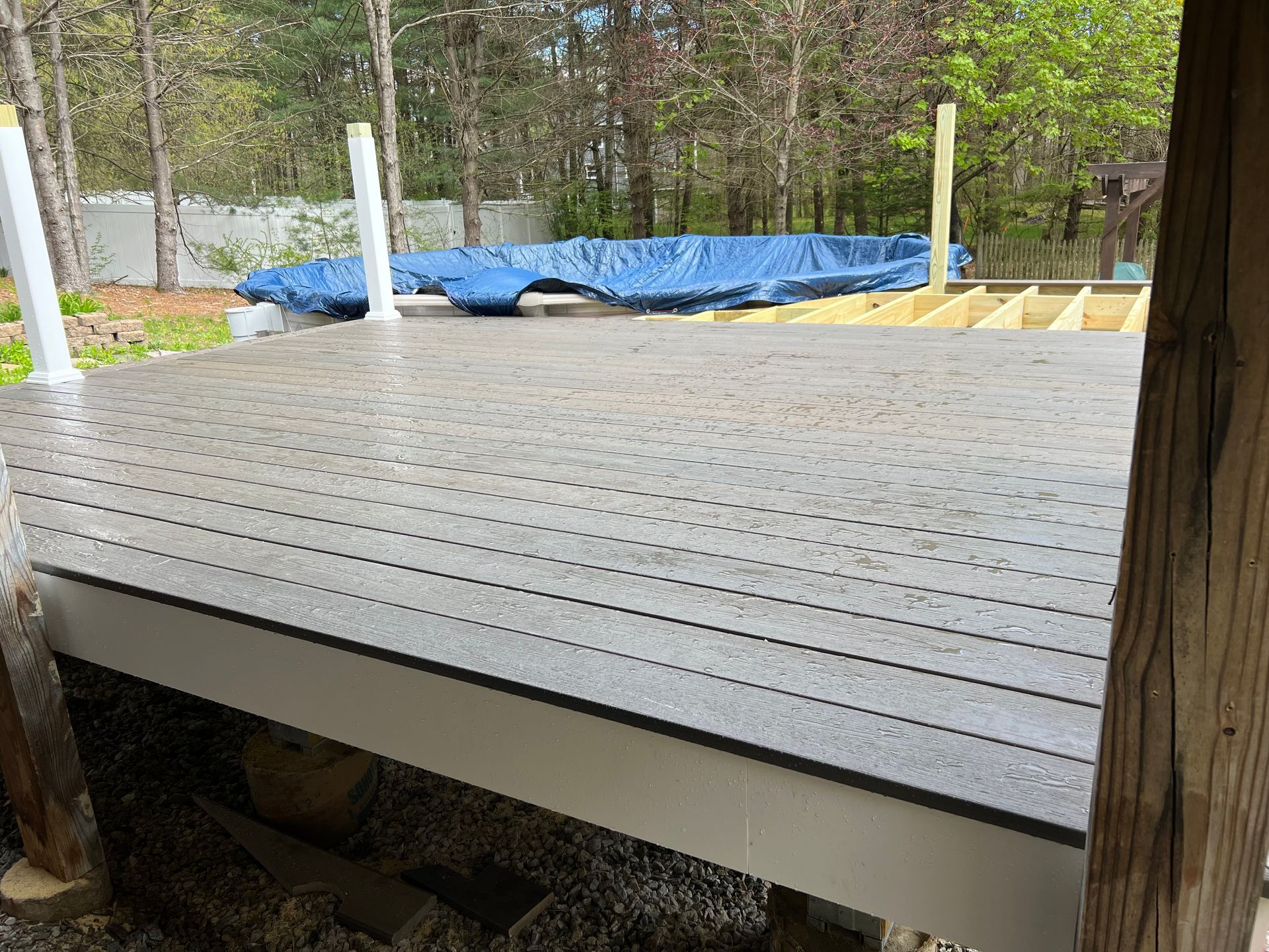 Gray composite deck with rain droplets. Blue tarp covers unfinished area; white railing posts.