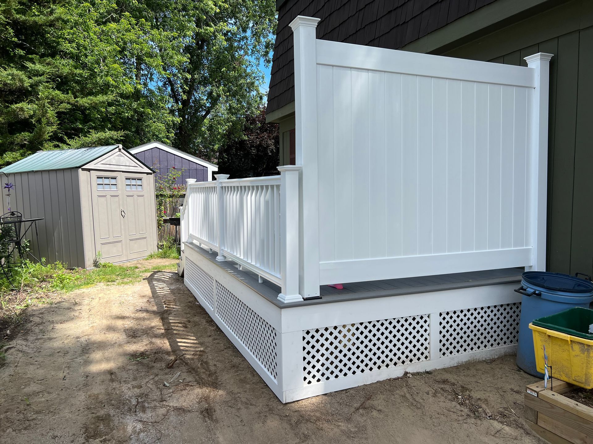White deck with lattice skirting, railing, and privacy screen next to a green building; two sheds in the background.