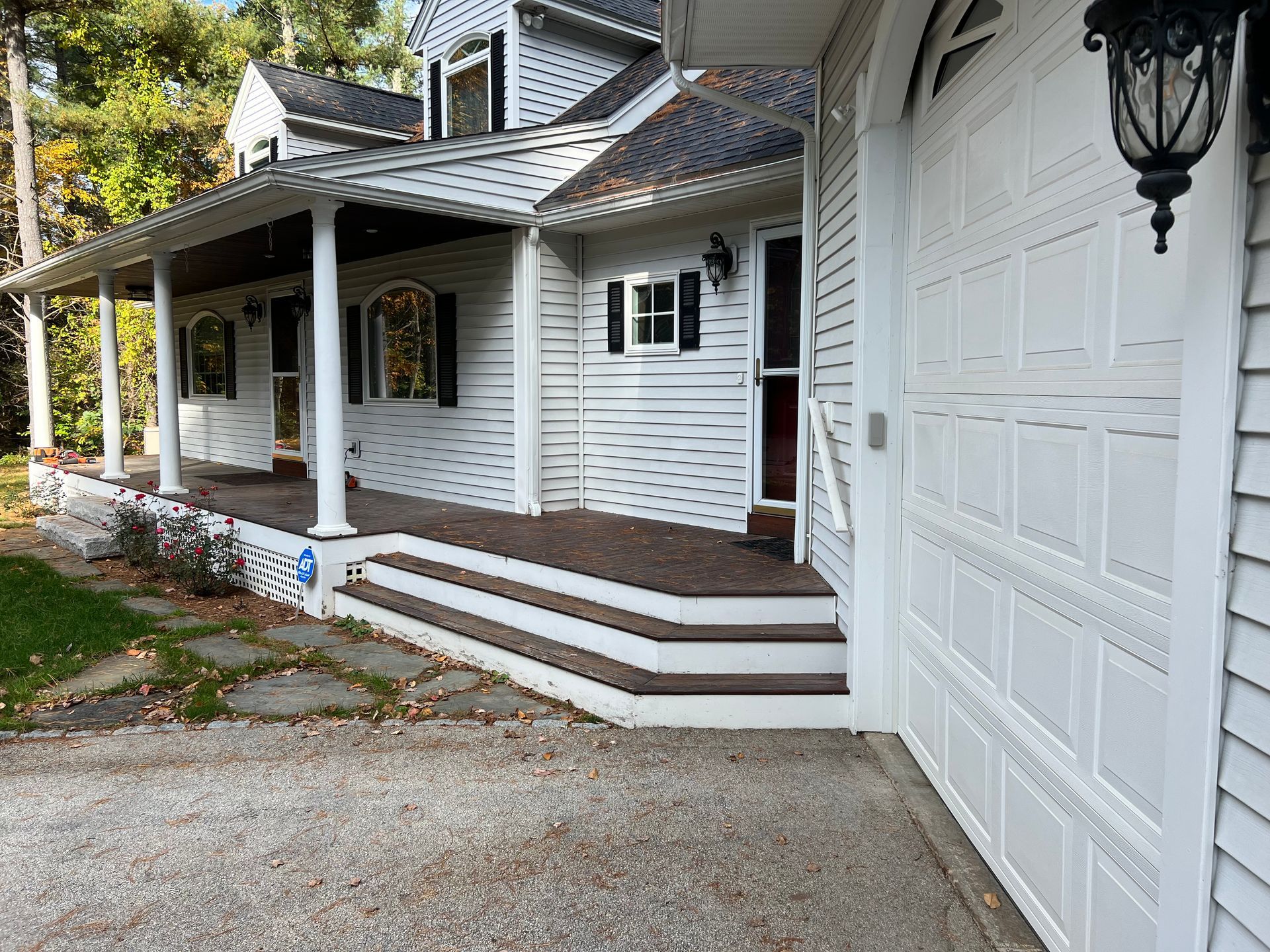 White house with covered porch, steps, and garage. Brown trim, gray driveway, and autumn leaves.