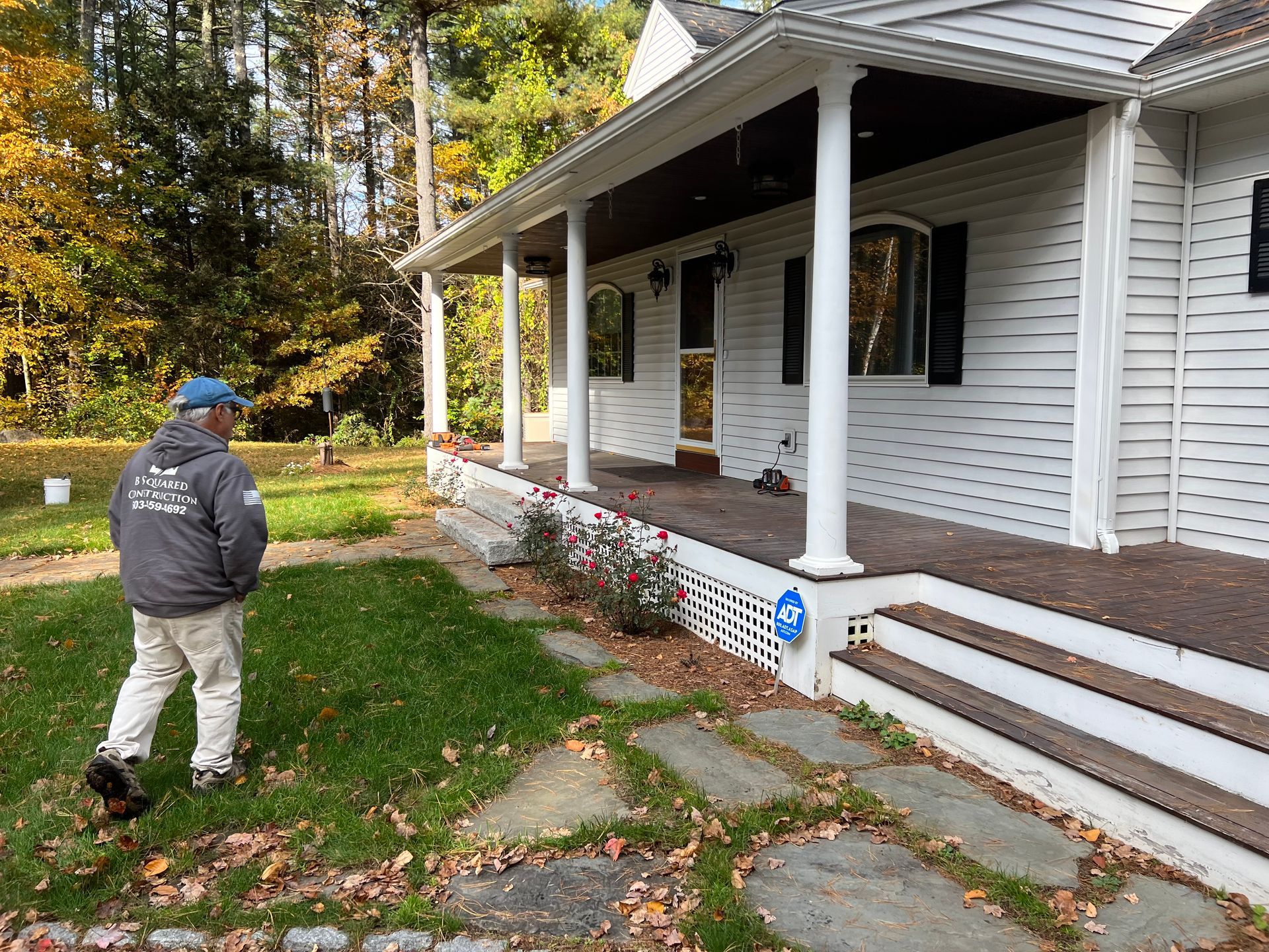 Man in front of white house with porch; stone path, green grass, fall foliage.