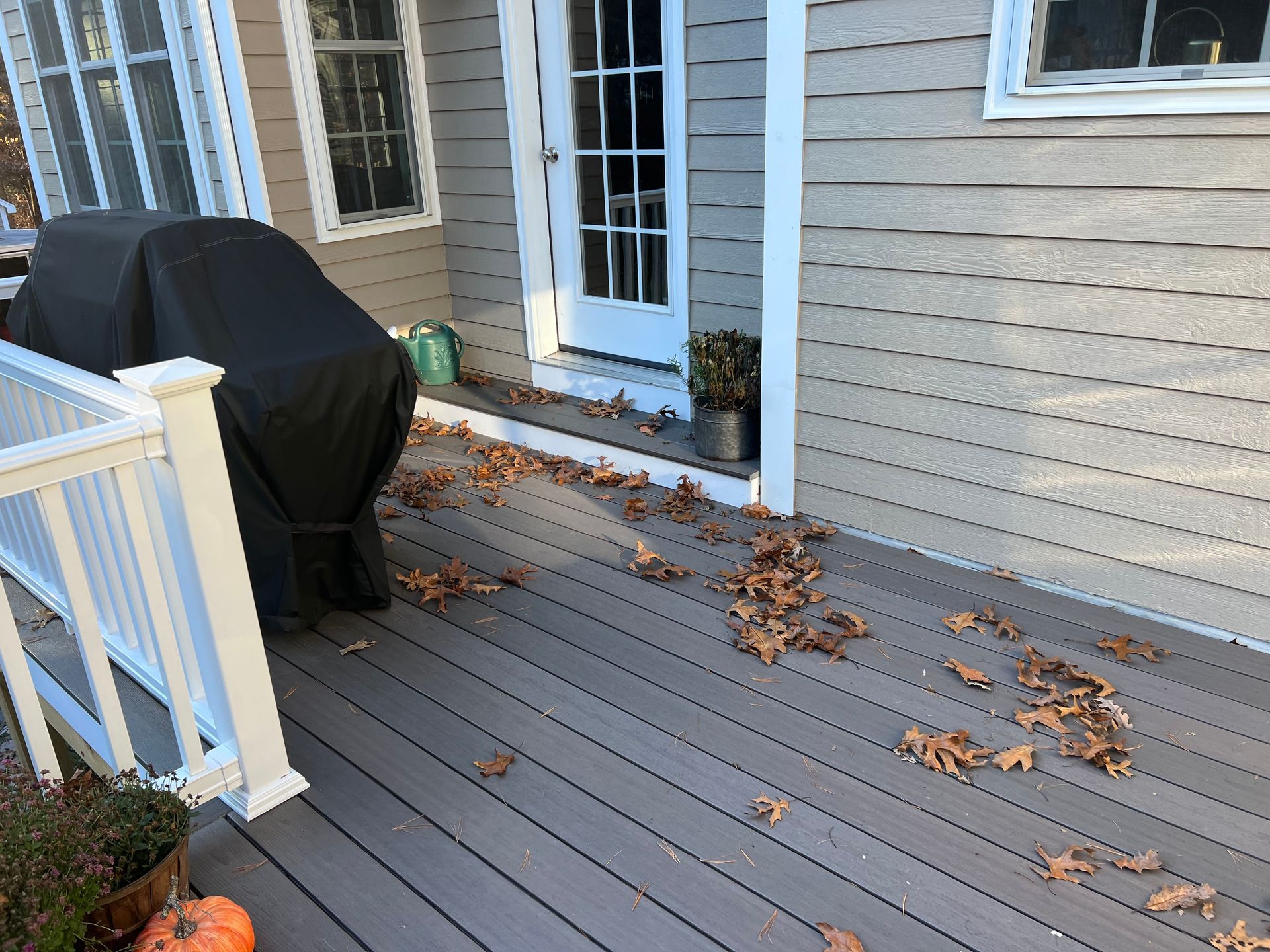 Deck with fallen leaves; house siding and white railing in the background; covered grill.