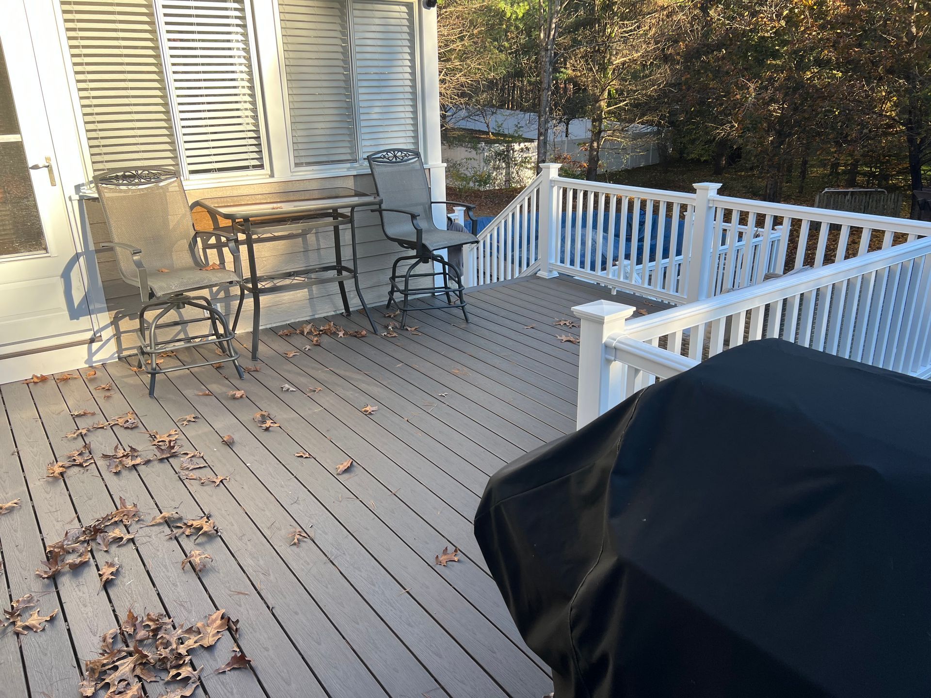 Deck with table and chairs, white railing, and grill, covered in fallen leaves, outdoors.