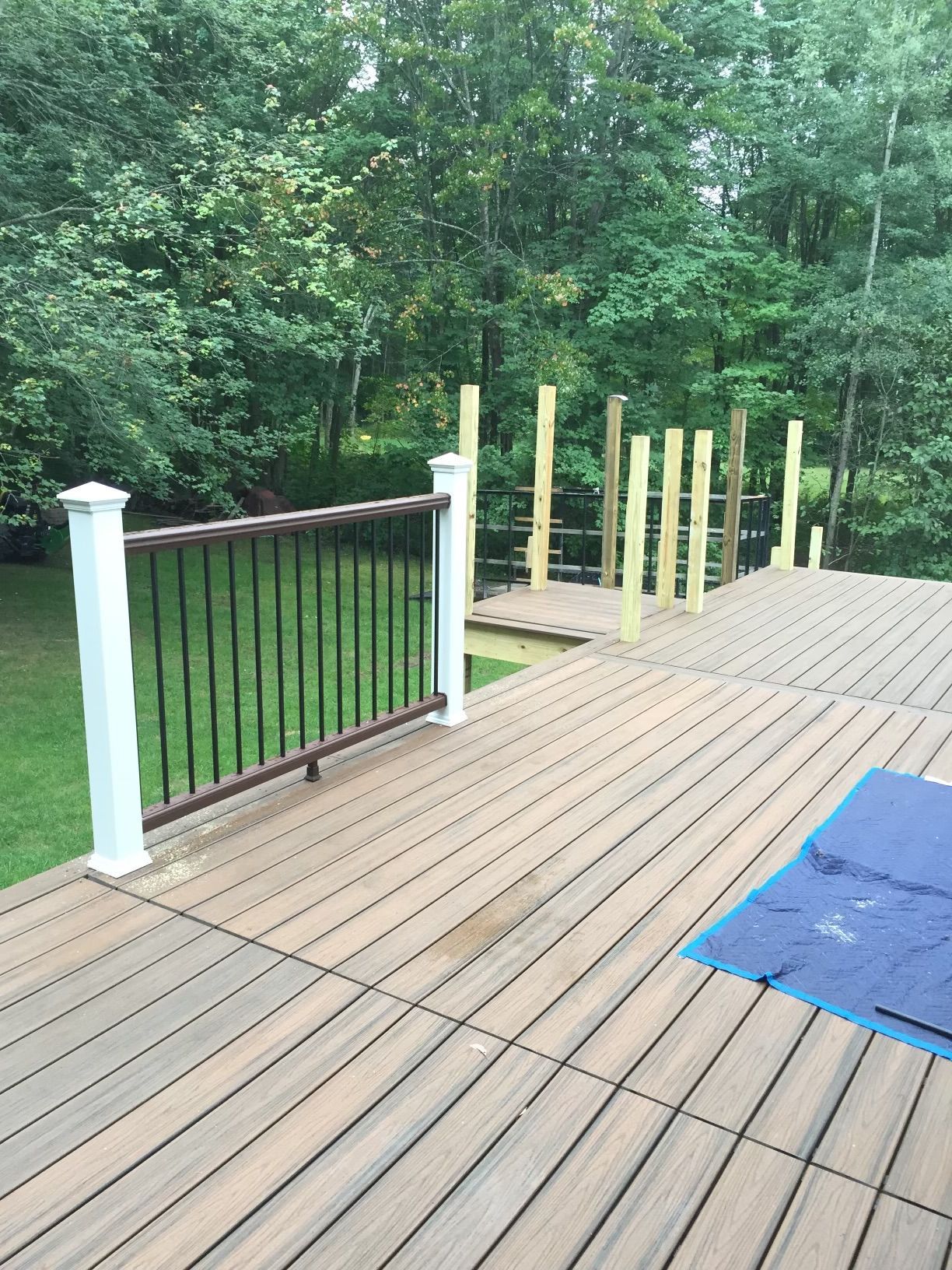 Deck with railing, unfinished posts, and a blue tarp on a wooden surface, surrounded by trees.