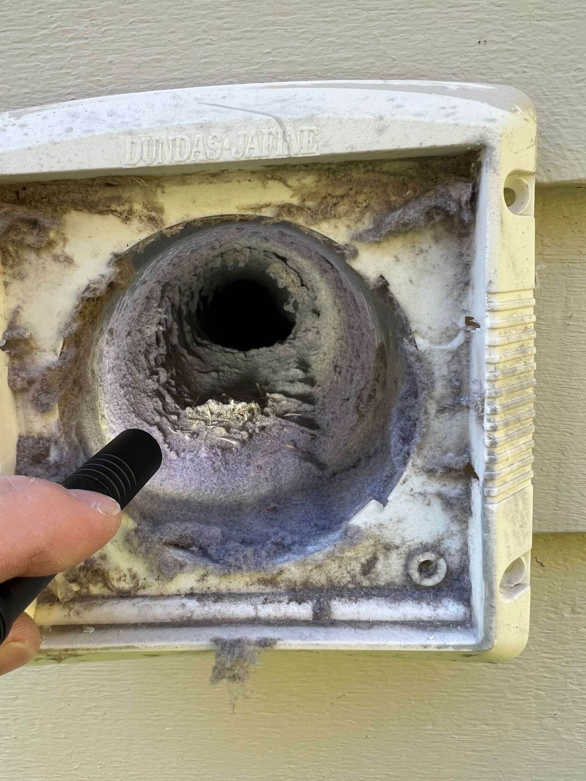 Clogged dryer vent covered in lint. A hand holds a tool inside. Set outdoors on beige siding.