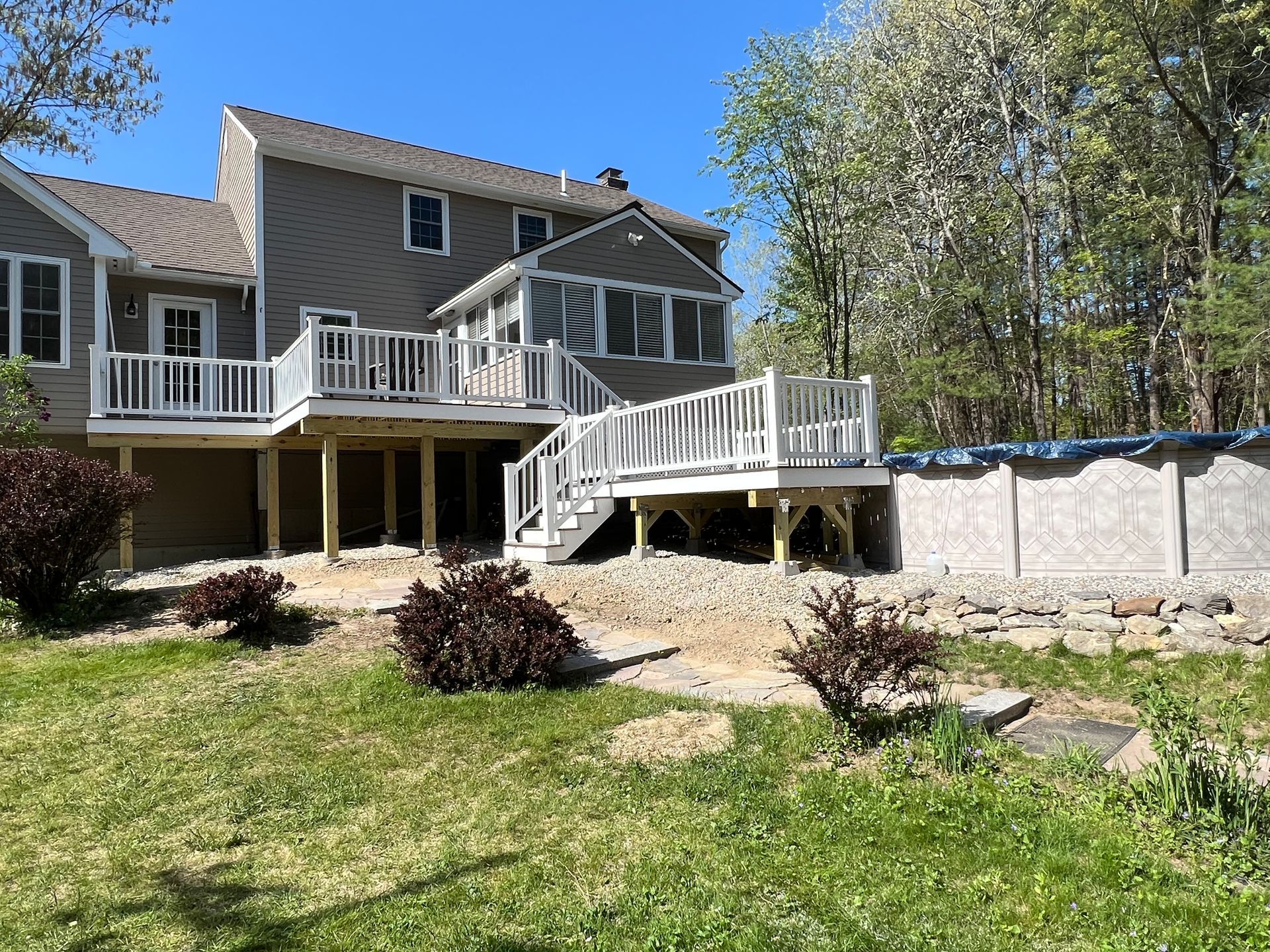 Backyard with house, deck, and above-ground pool. Green grass, gravel, and trees surround the deck.