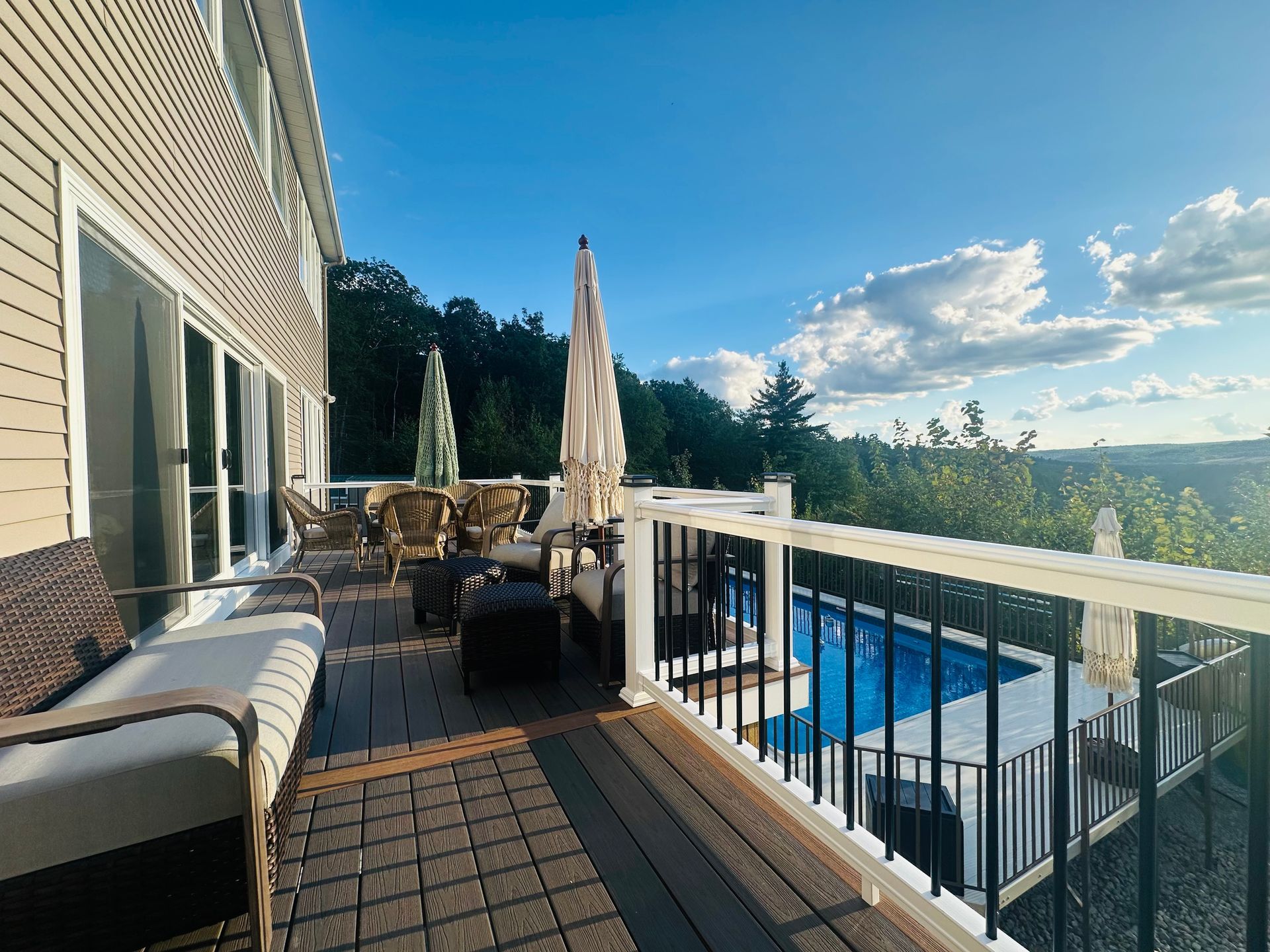 Deck overlooking pool, blue sky, trees in the background. Furnished patio with seating, umbrellas, and wooden deck.