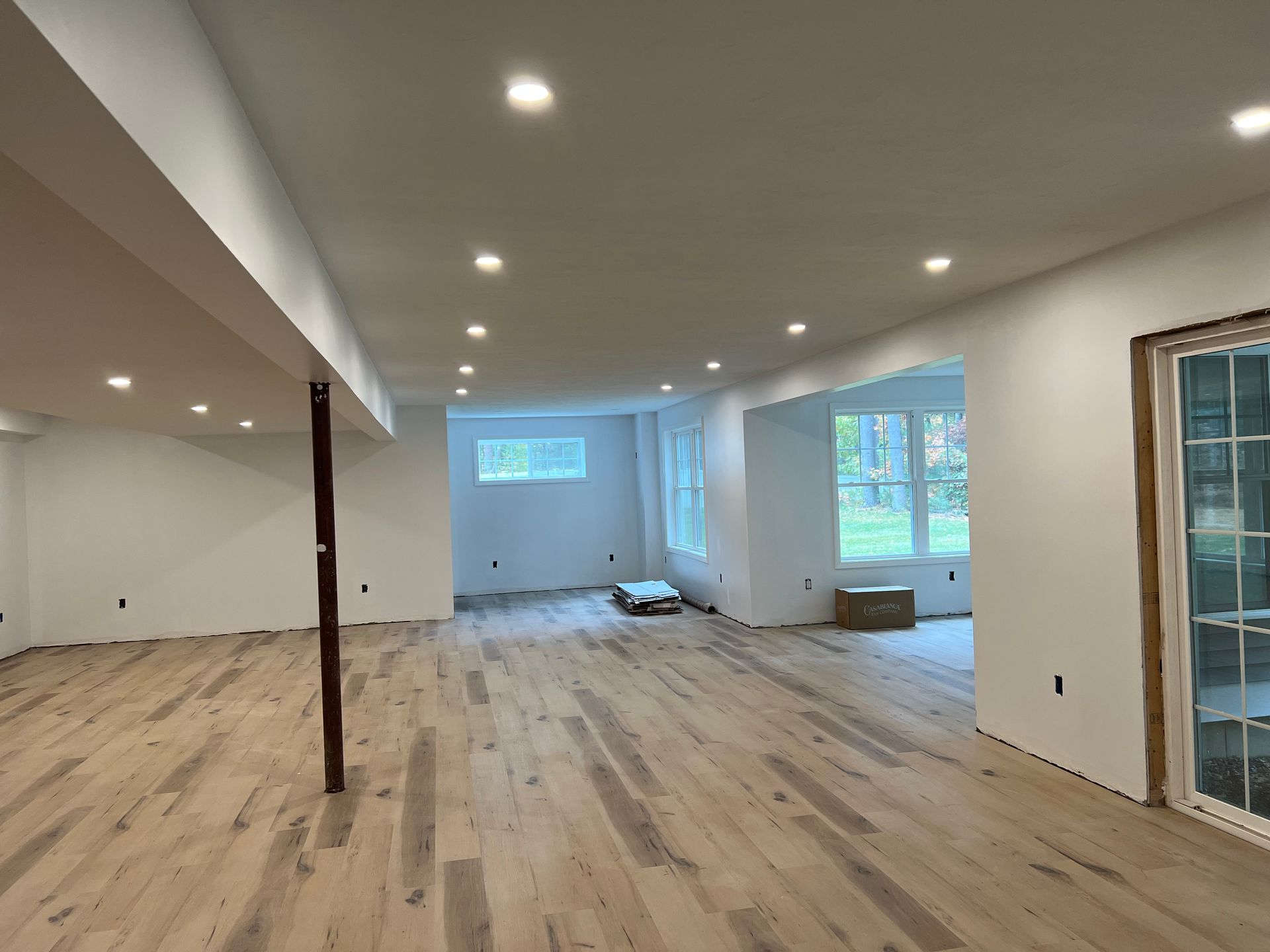 Empty room with light wood floor, white walls, recessed lighting, and windows.