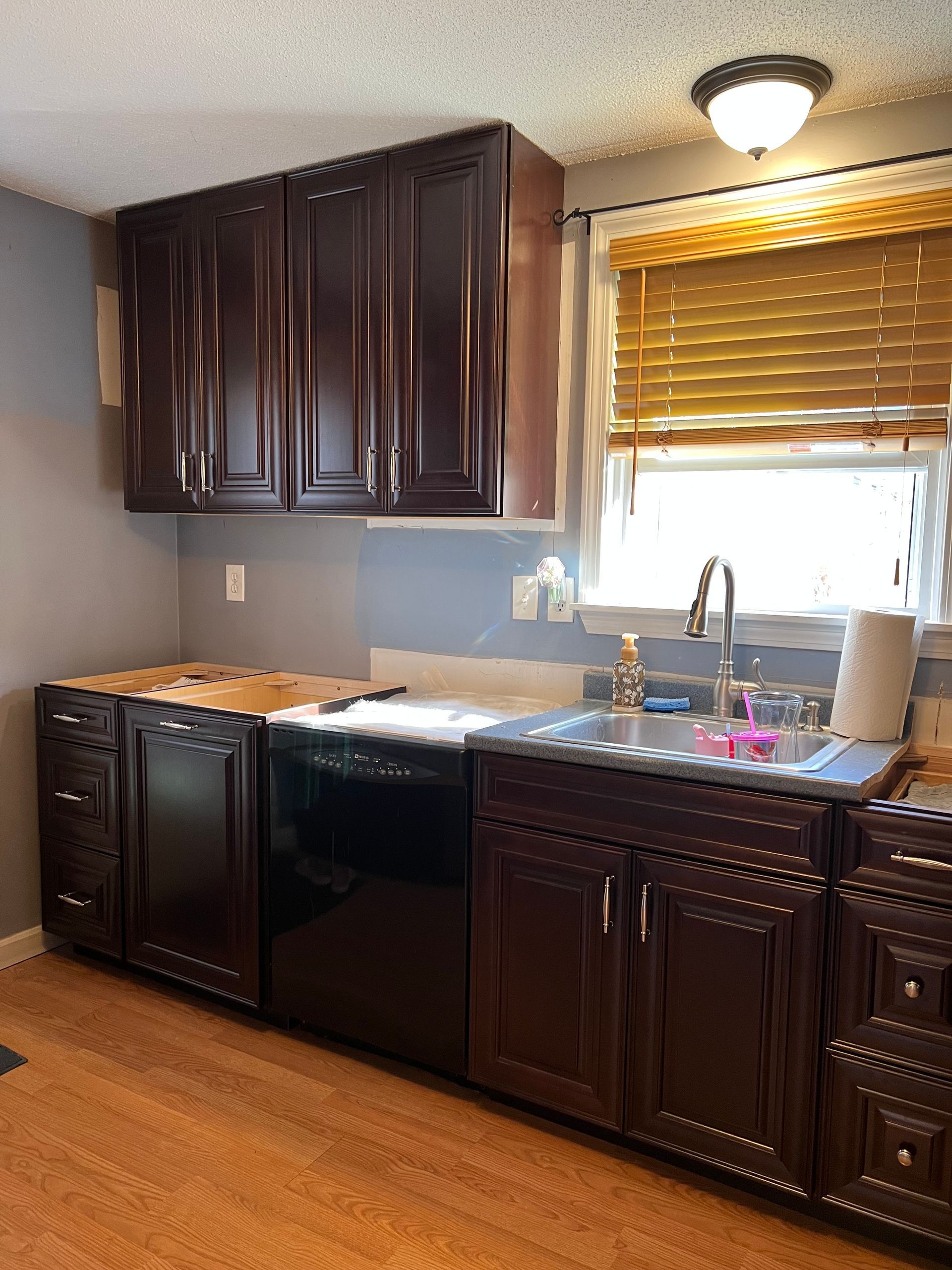Kitchen renovation in progress. Dark cabinets installed, light blue wall. Dishwasher and sink visible.