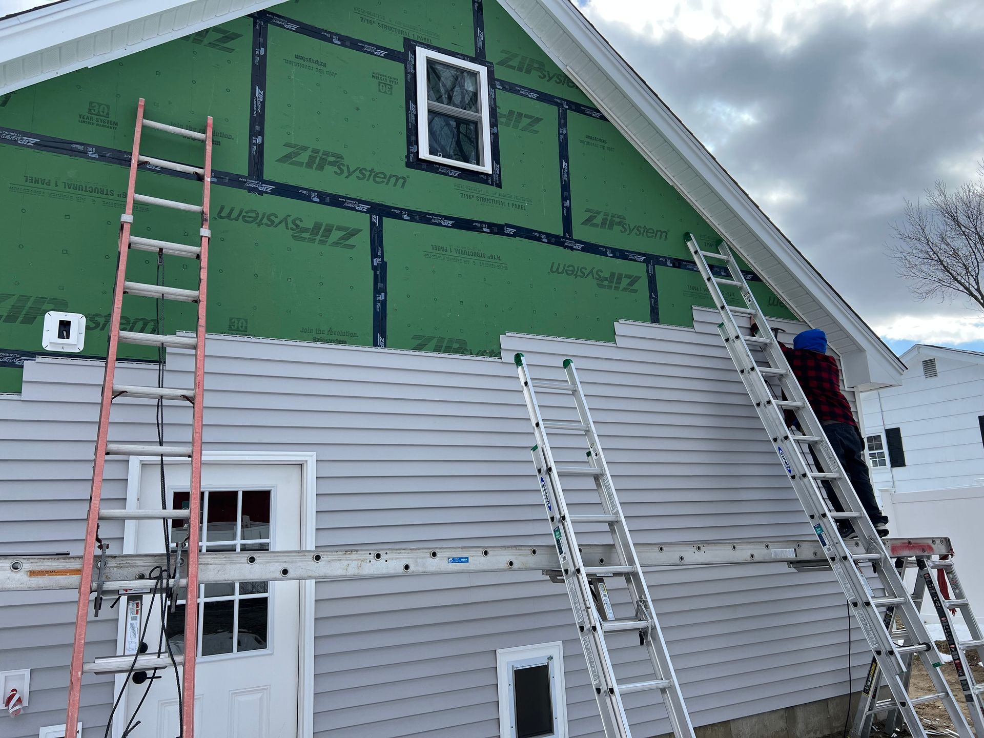 Workers install siding on a house. Ladders are propped against the wall. Green sheathing is visible.