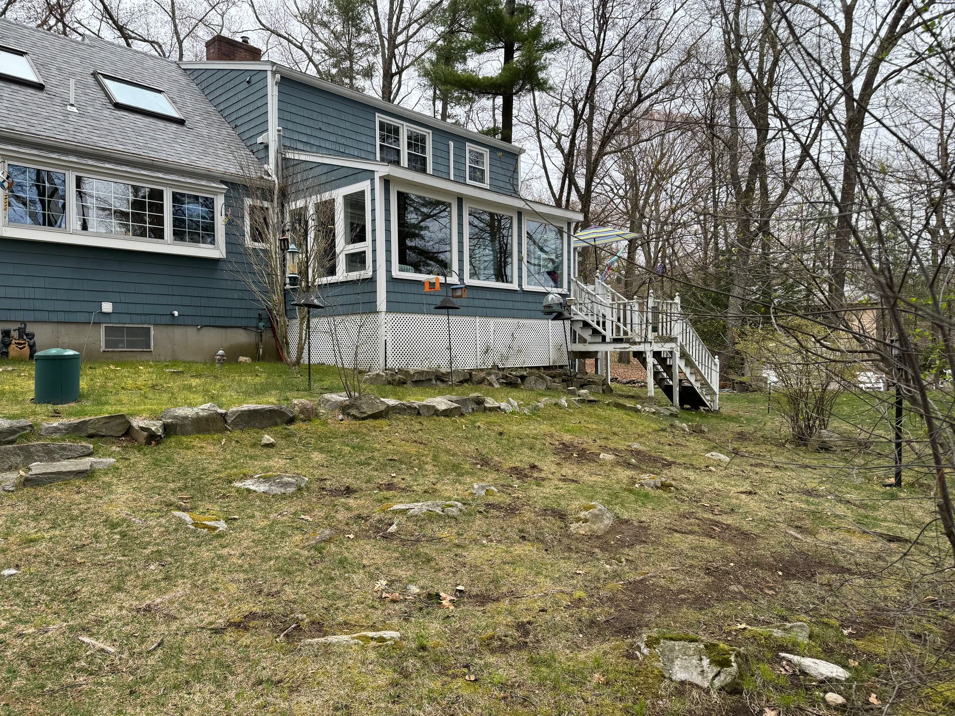 A blue house with many windows and a deck sits on a grassy hillside in a wooded area.