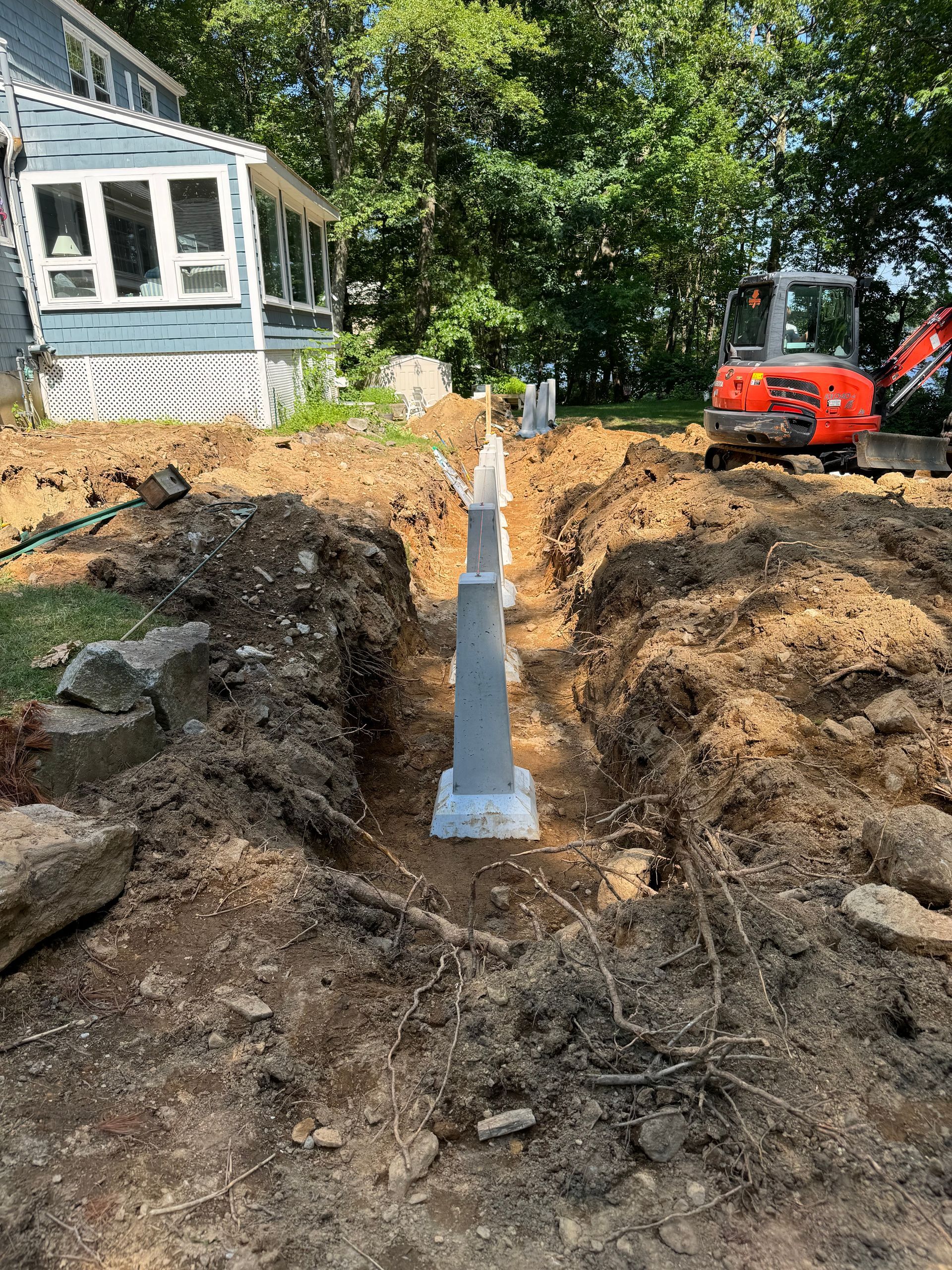 Construction site with concrete posts in a trench, a mini excavator, and a light blue house.