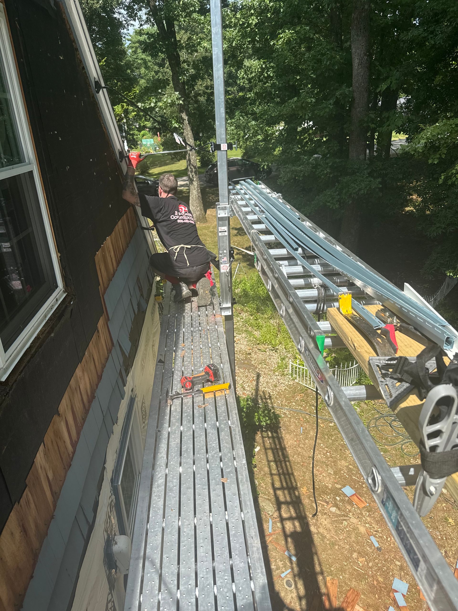 Scaffolding alongside a house, tools visible. A worker stands on the platform near trees.