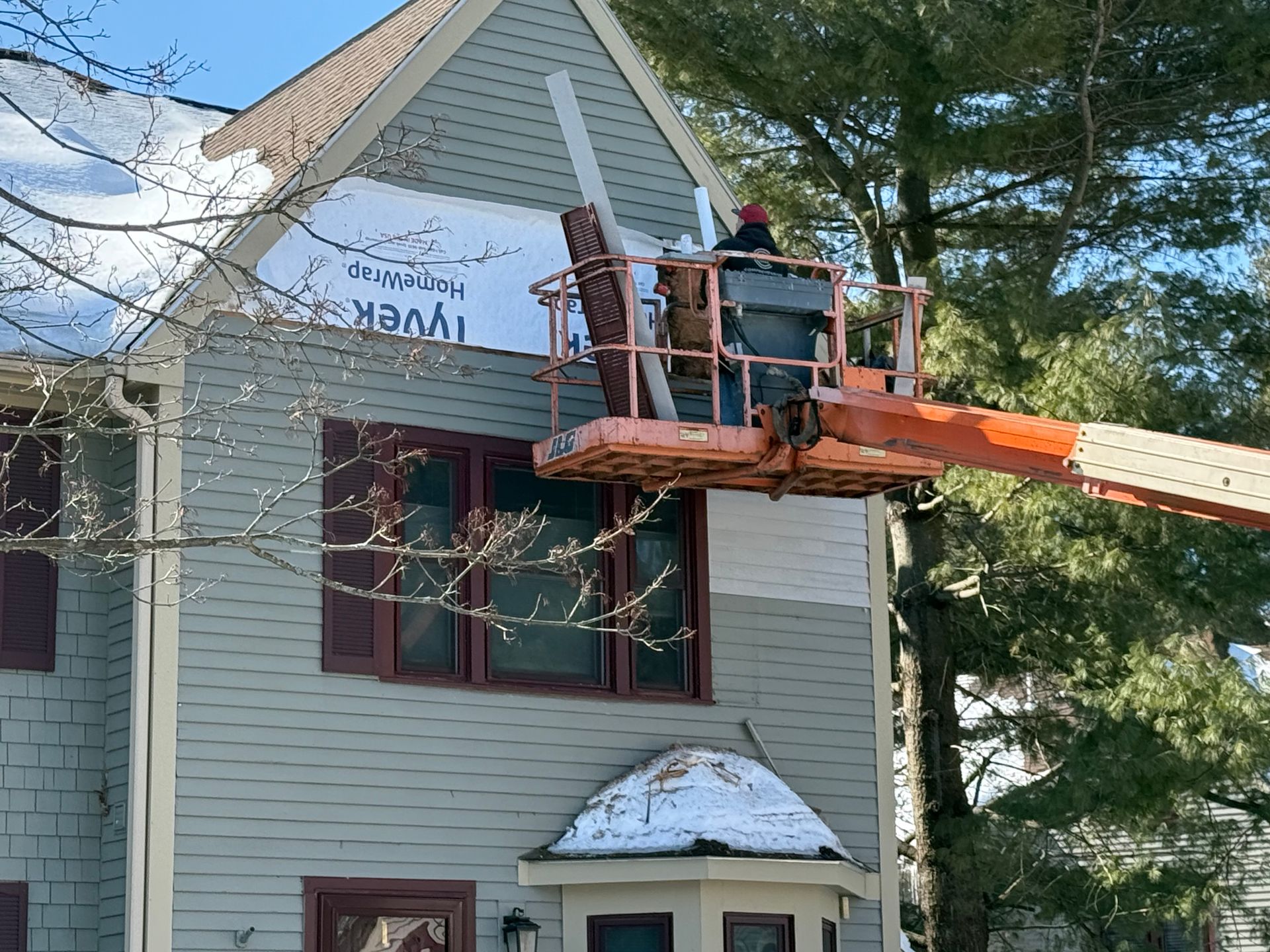 Man in lift installing siding on a two-story house, white Tyvek exposed, snow on the roof.