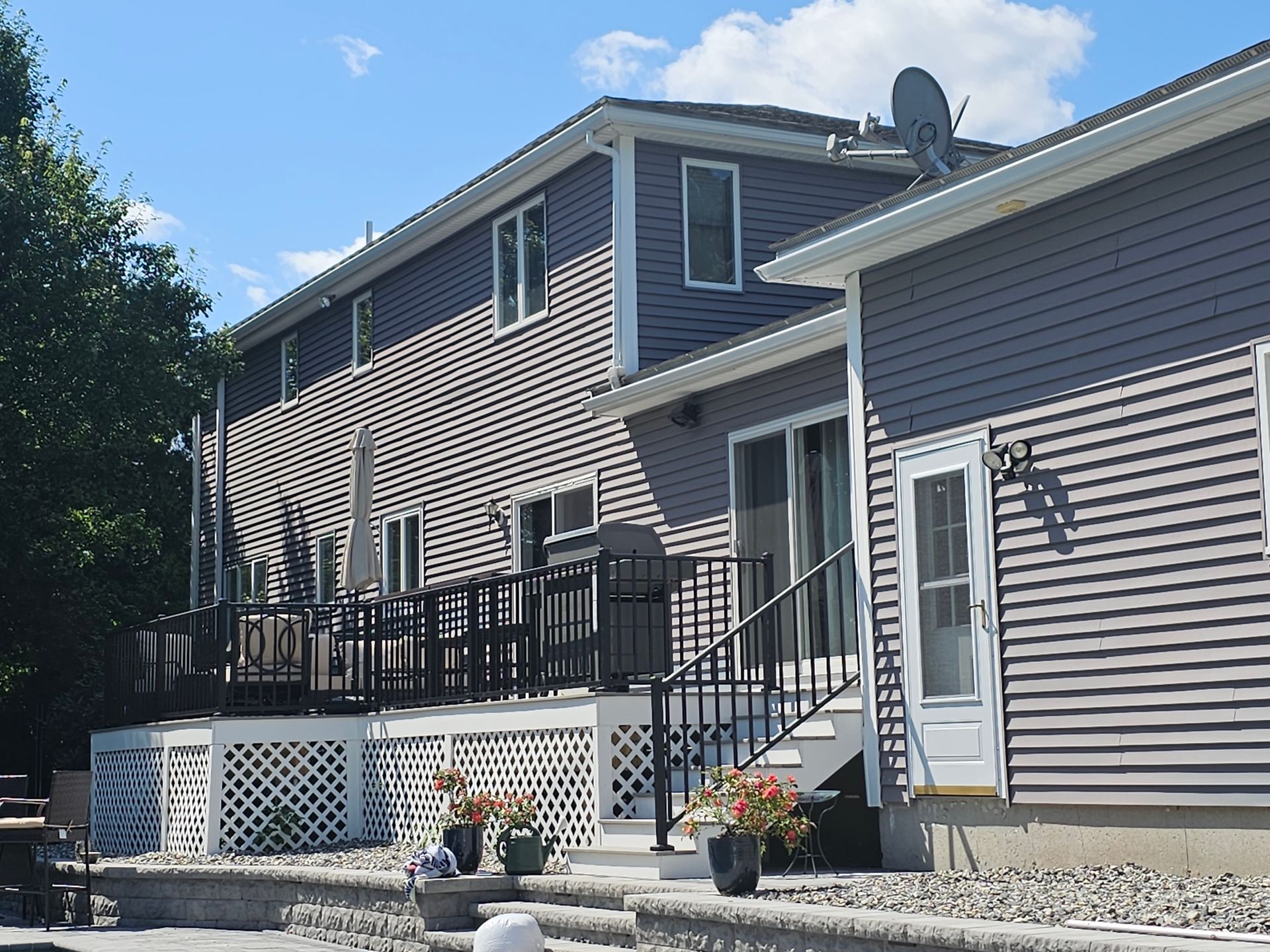 Two-story gray house with deck, stairs, and outdoor seating; sunny day.