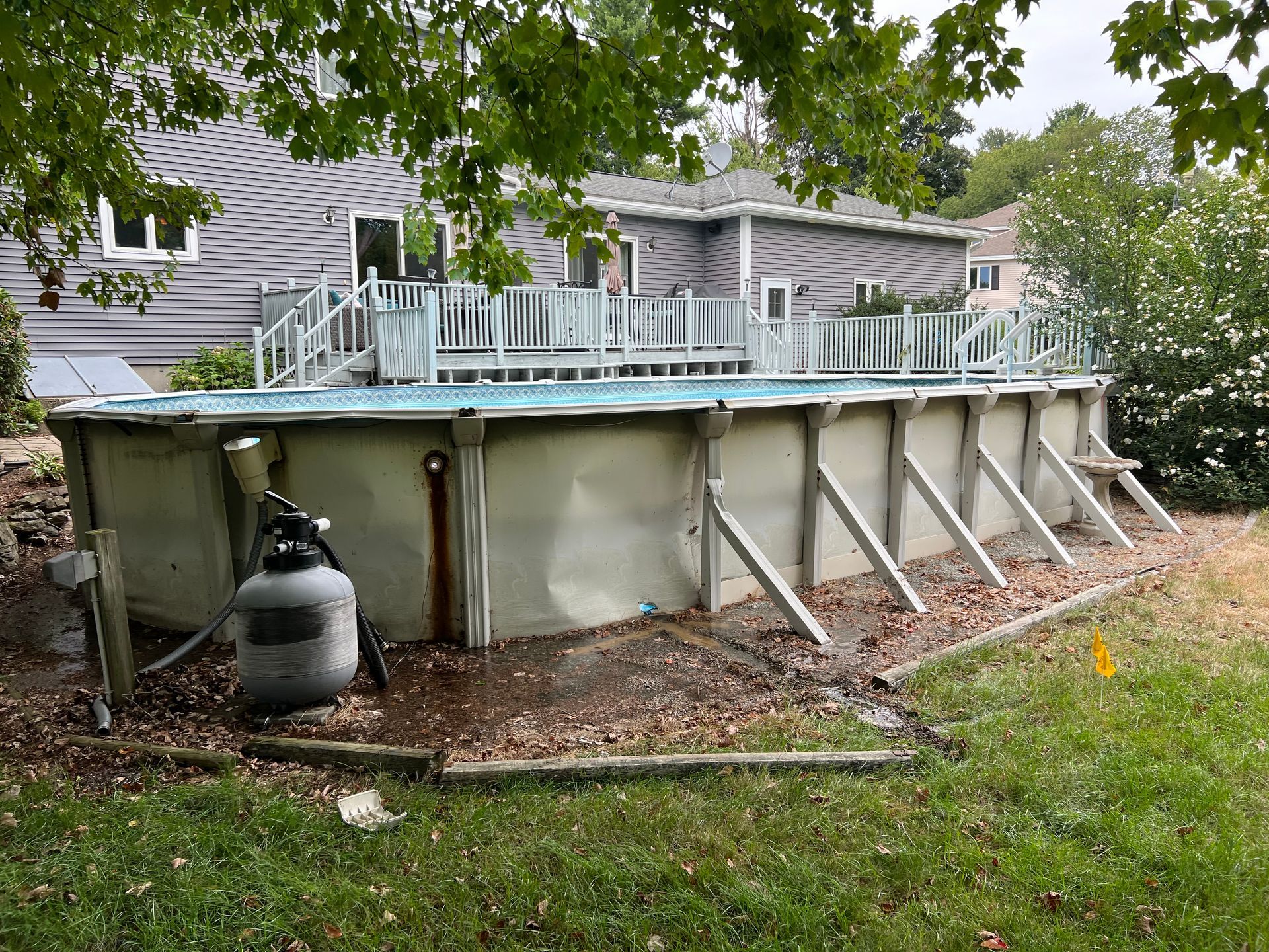 An above-ground pool in a backyard. The pool is surrounded by fencing and sits on a platform of dirt and wood.