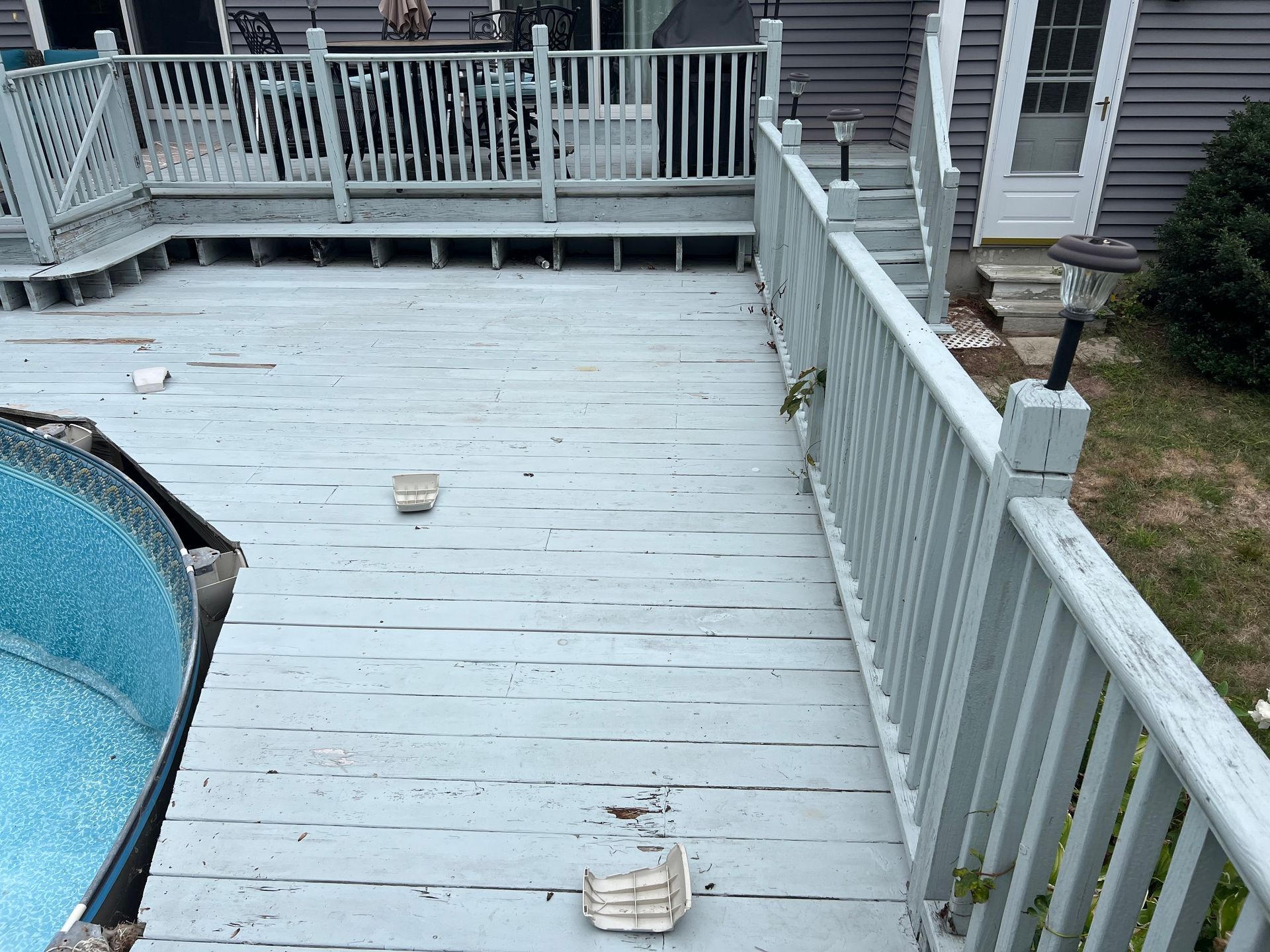 Gray wooden deck with missing planks and railing, near an above-ground pool and a house door.