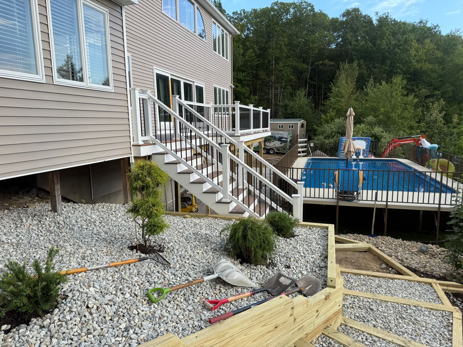 House with deck, stairs, and pool, landscaping with gravel, trees, and retaining walls.