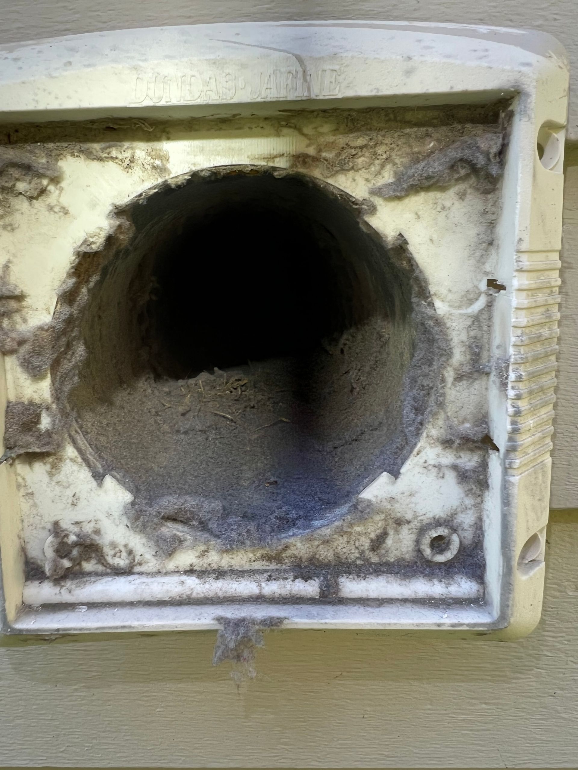 Close-up of a dirty dryer vent, clogged with lint, on a light-colored exterior wall.