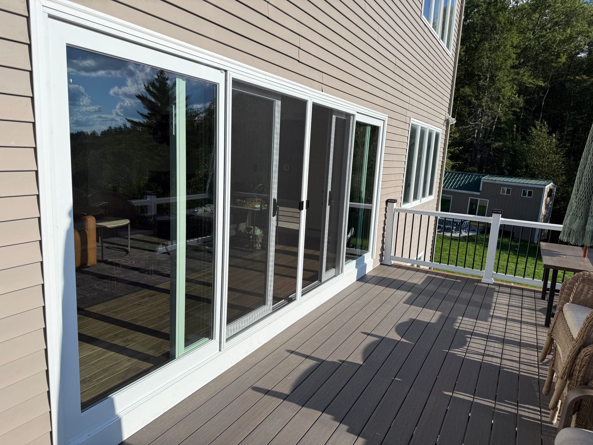 Deck with sliding glass doors; beige siding and gray deck. White frame doors reflect trees and deck furniture.