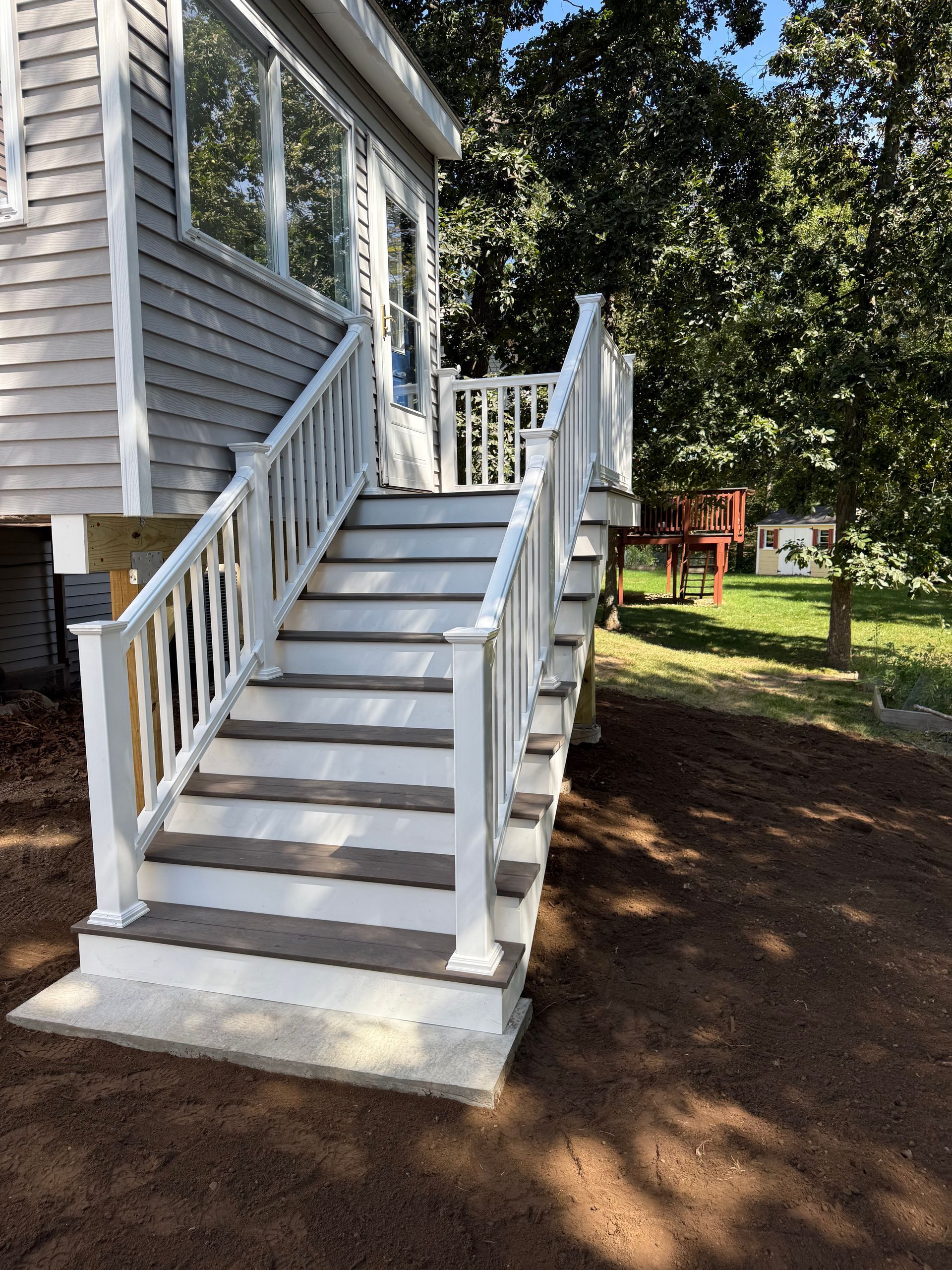Exterior stairs with white railings leading to a house, set in a yard with trees.