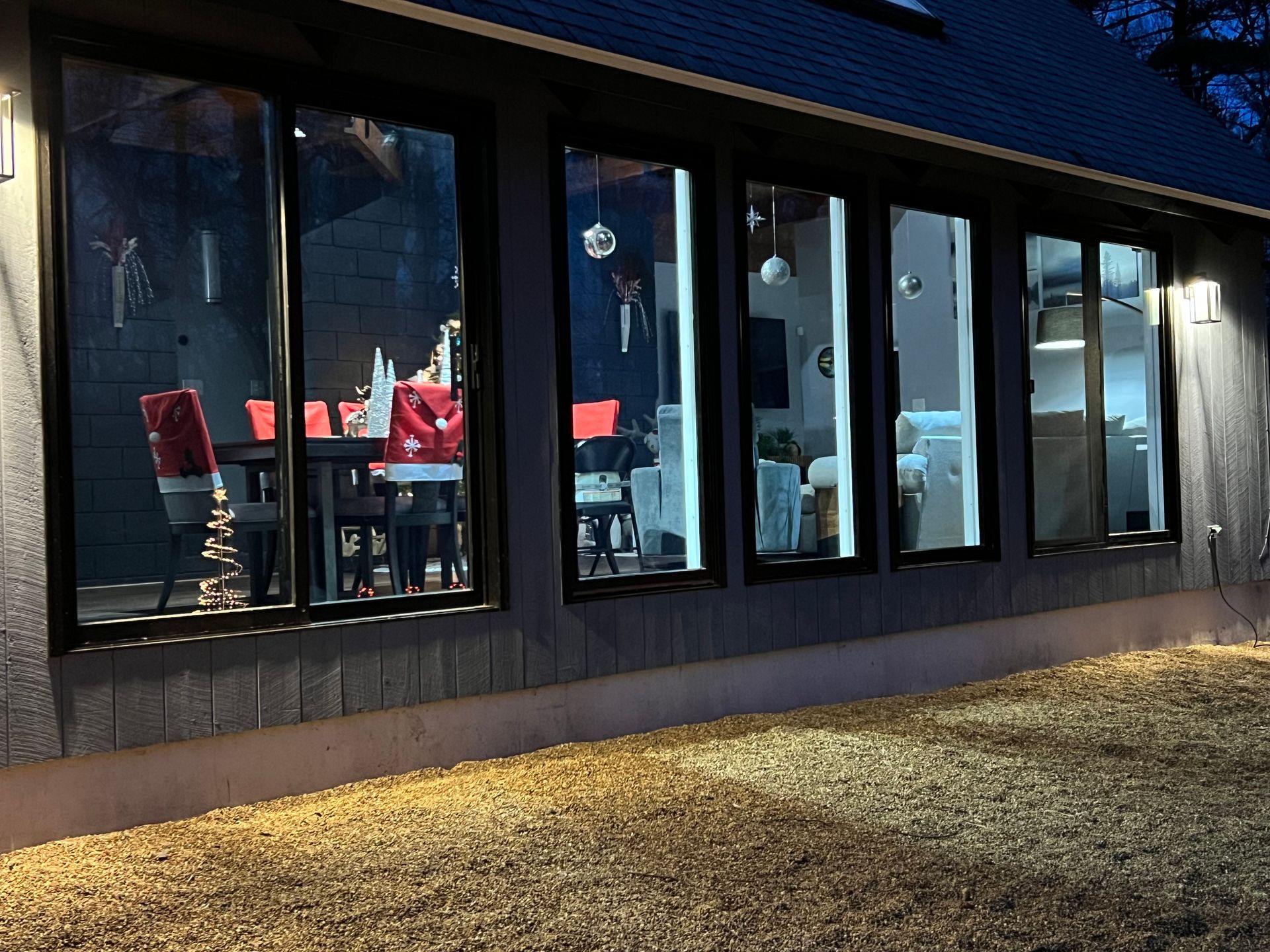 Exterior view of a house with large windows reflecting a dining room scene, illuminated by outdoor lights.