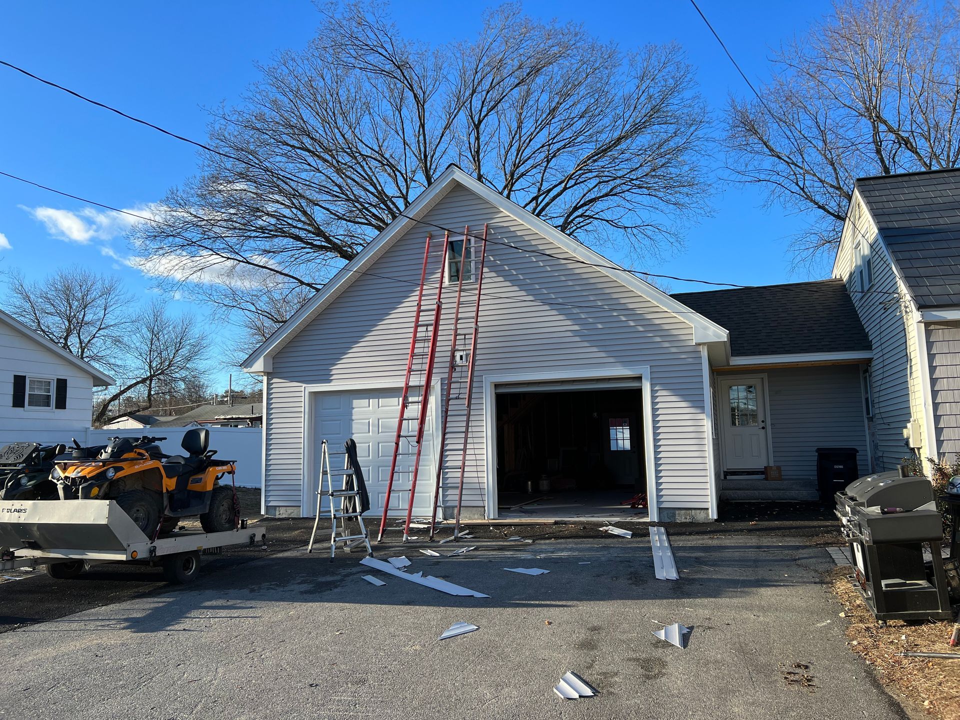 Garage being built, light grey siding, open door, red ladder, man on ATV, debris on ground.