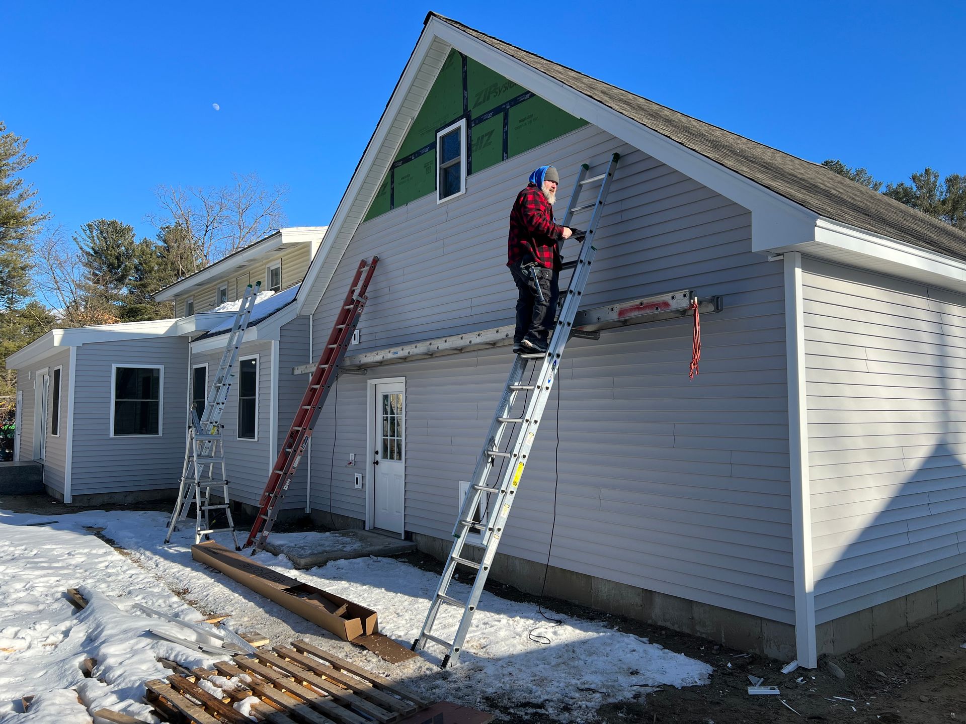 Man on ladder installing siding on a house exterior. Snow on ground.