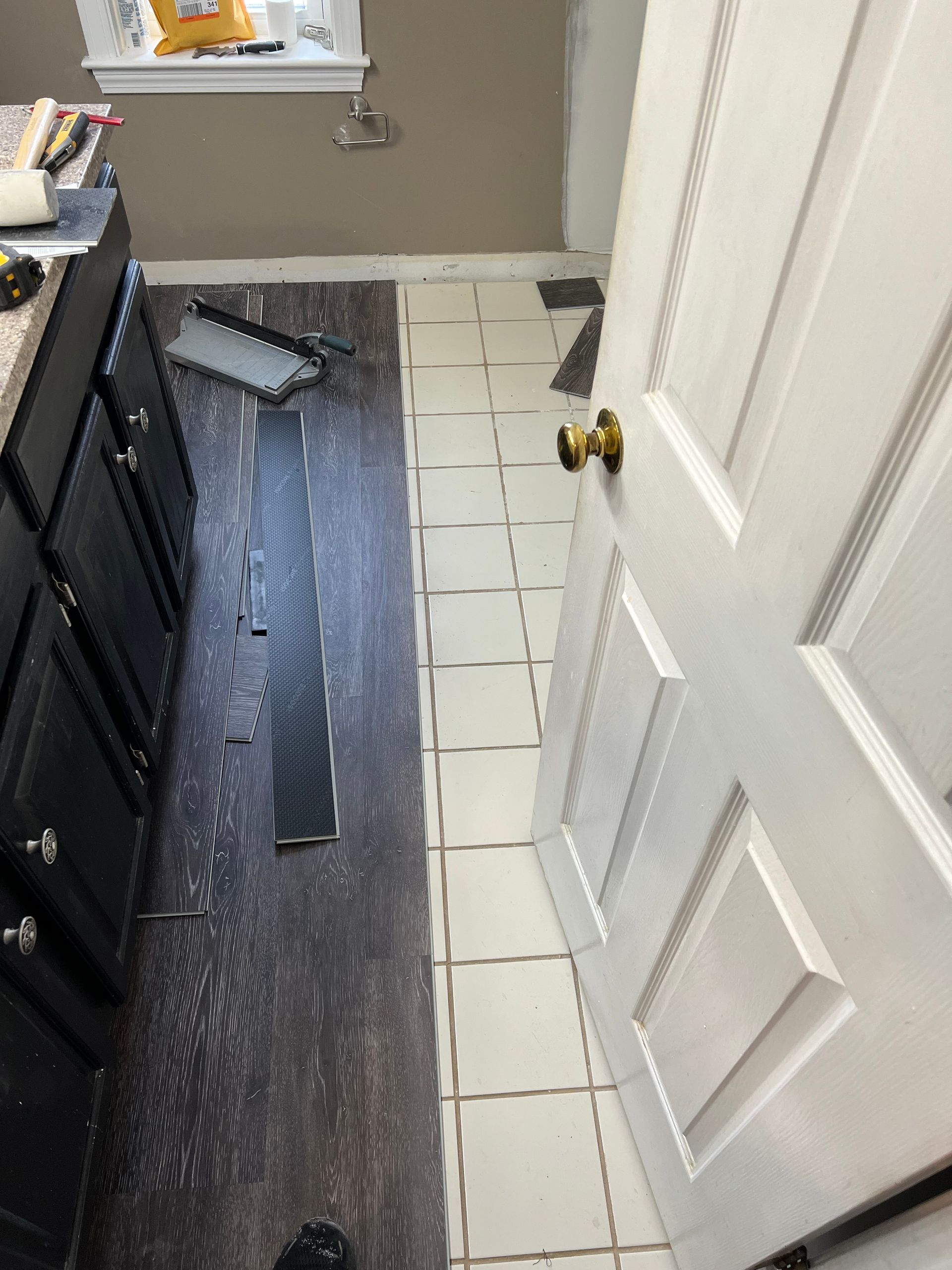 Dark wood-look flooring installation beside white tile floor; dark cabinets and a partially open white door are visible.