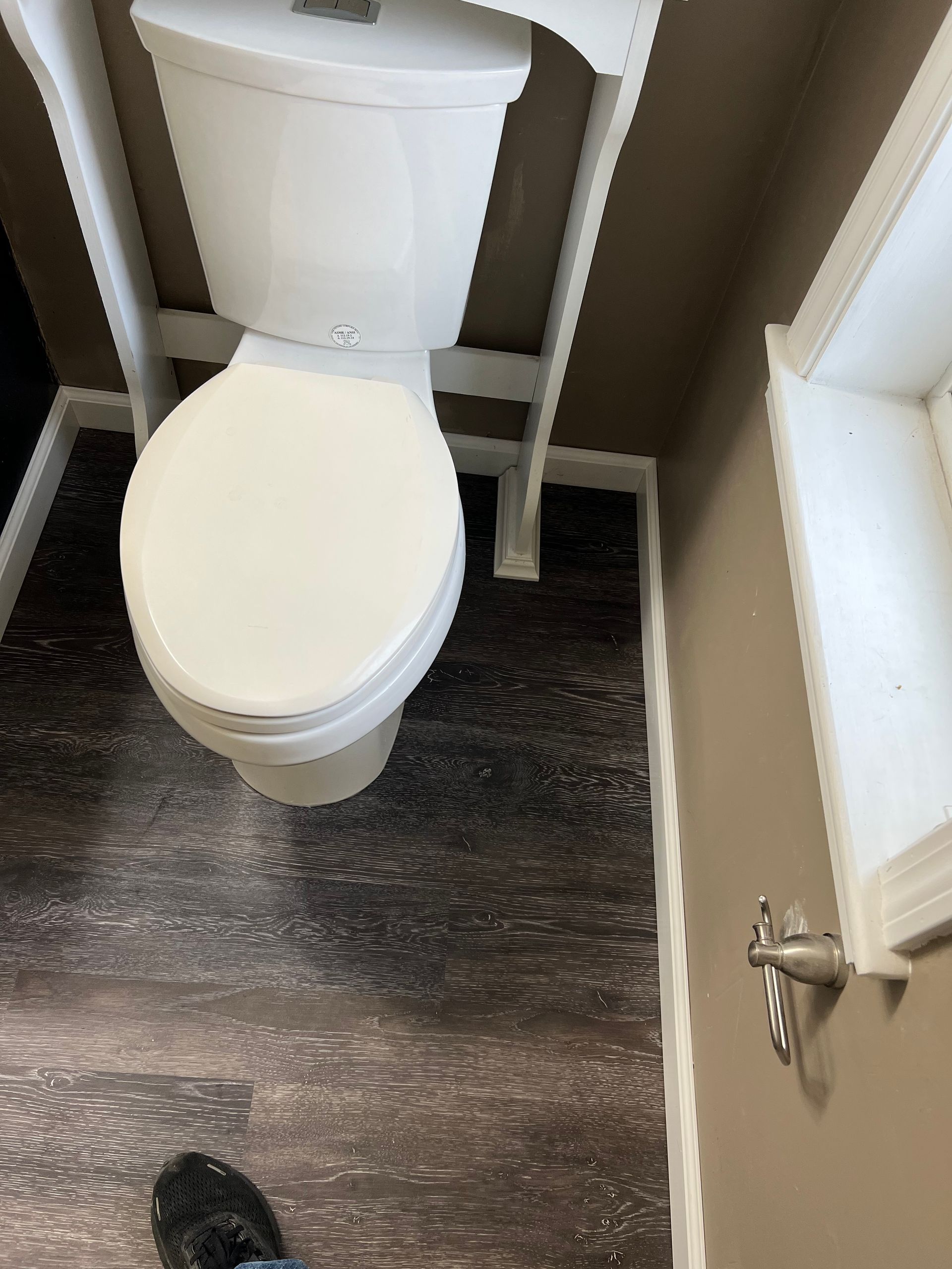 Toilet in a small bathroom, white seat, dark wood-look floor, tan walls, and a window.