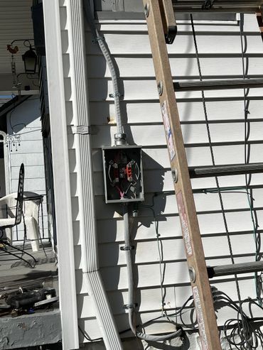 Electrical box mounted on a white house exterior with conduit and ladder nearby.