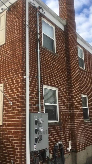 Exterior view of a brick building with electrical conduit running vertically. A gray electrical box is mounted on the wall.
