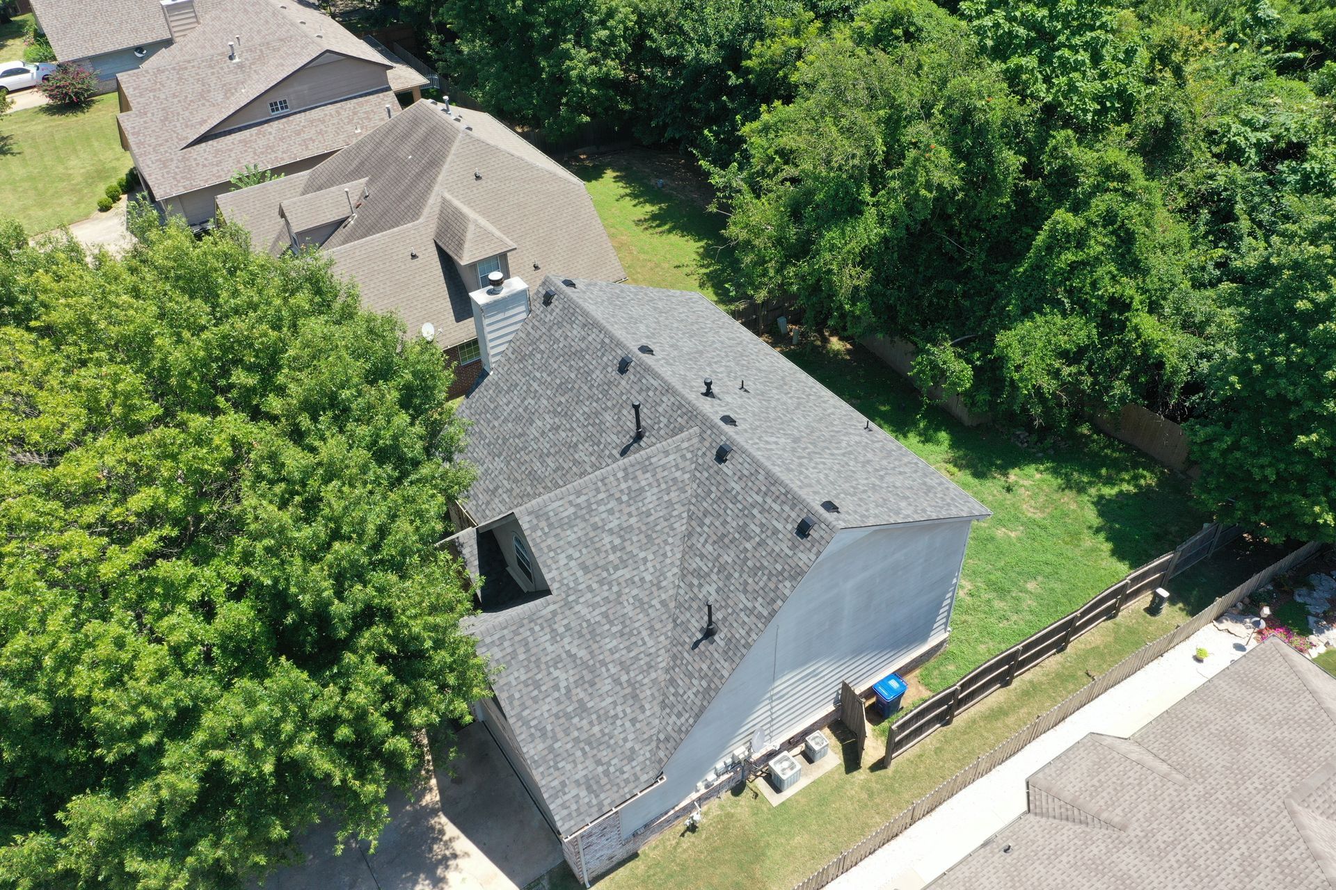 Aerial view of a gray-roofed house with surrounding trees and neighboring homes on a sunny day