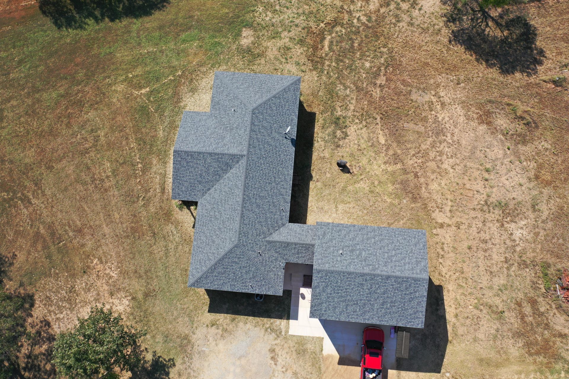 Aerial view of a gray-roofed house with a driveway and a red car, surrounded by dry grass.