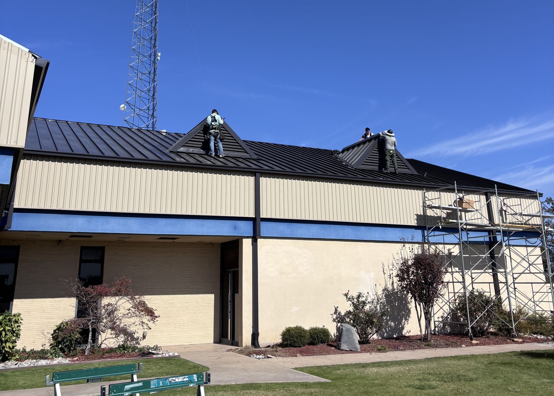 Large beige building with black metal roof under a clear blue sky, with scaffolding on the right side.