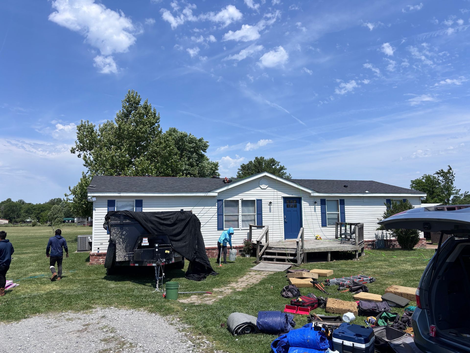 People unloading belongings outside a white mobile home on a grassy lot under a blue sky