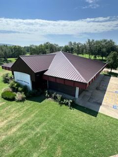 Aerial view of a dark brown building with a red metal roof beside a large green lawn and trees