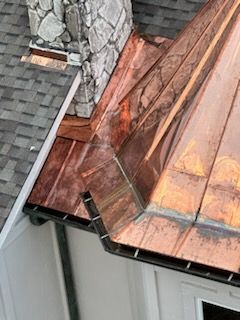 Copper roof valley and flashing beside a gray shingle roof and stone chimney.