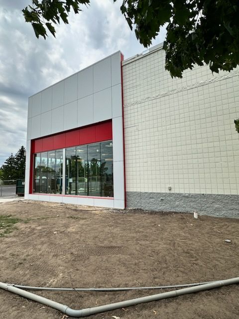 New building exterior with glass entrance, red awning, and unfinished dirt foreground under cloudy sky