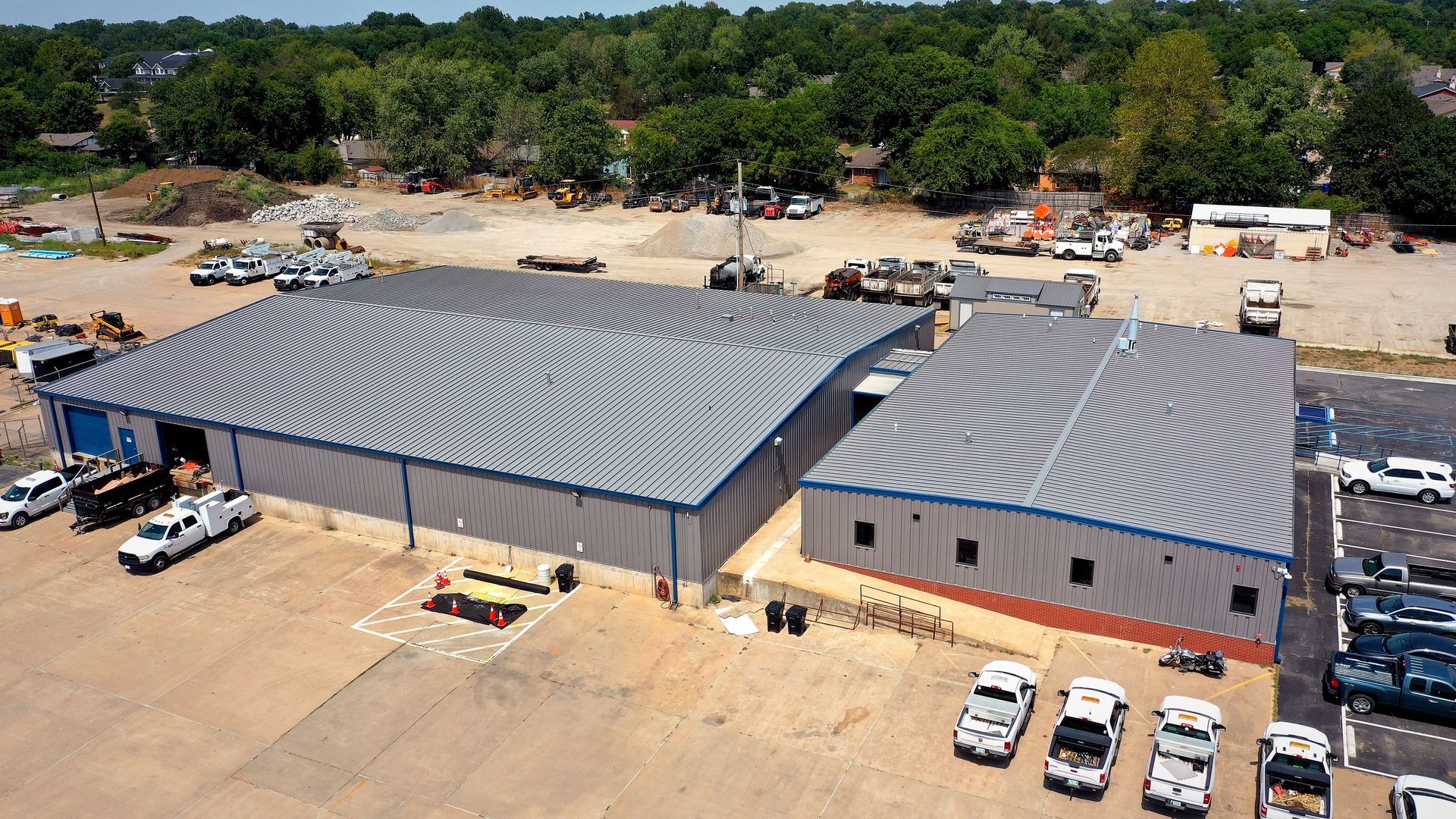 Aerial view of two gray-roofed buildings with parked cars in a dusty lot beside trees.