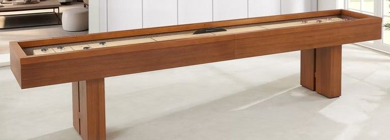 A long, brown wooden shuffleboard table on a light-colored floor, against a blurred interior.